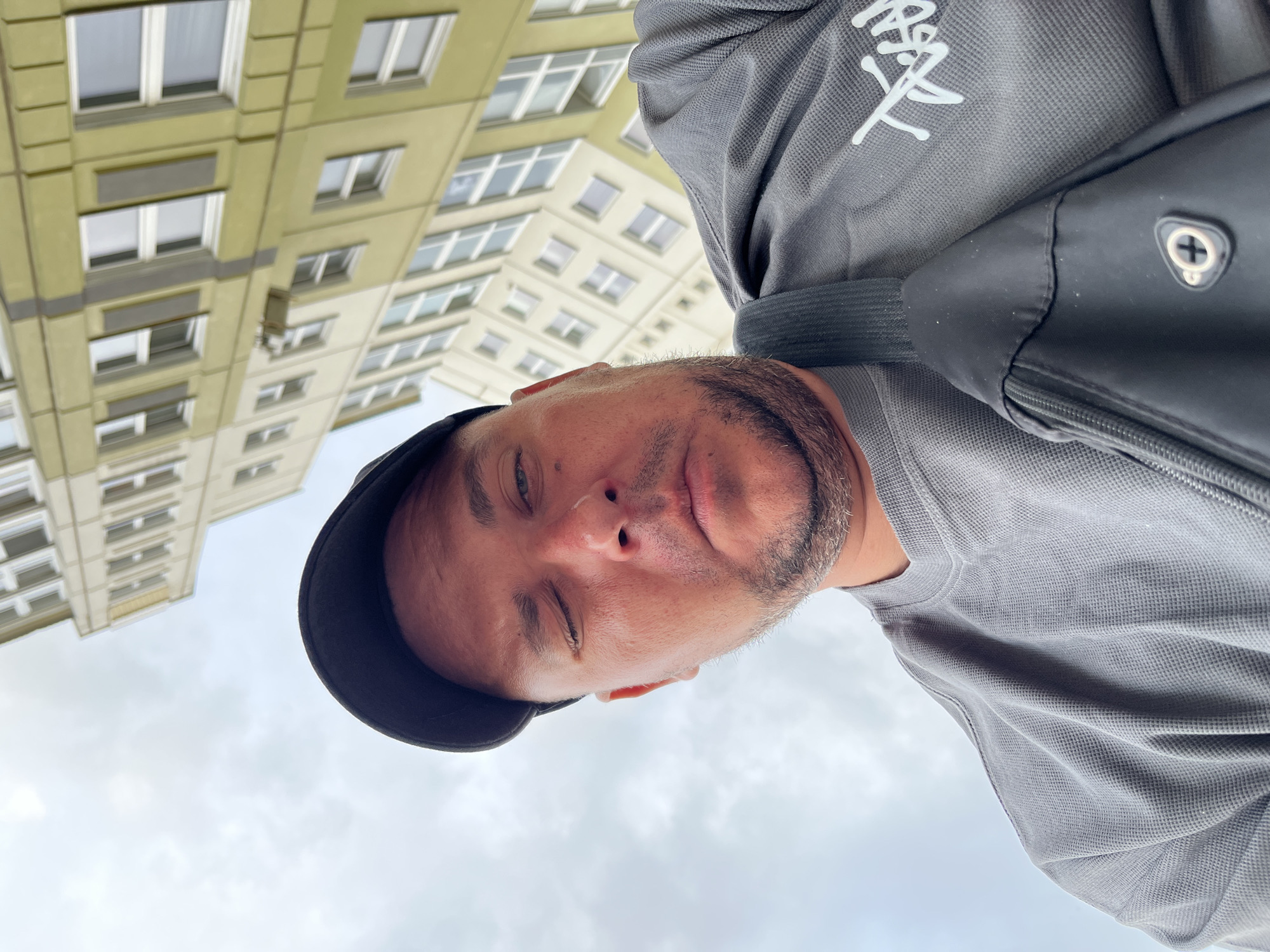 A male individual is captured in a low-angle shot, looking directly at the viewer. He has light skin, short stubble on his face, and wears a dark baseball cap. His left eye (viewer's right) is closed, creating a winking expression, while his right eye is partially open. He is dressed in a grey, textured, long-sleeved shirt with a white, stylized graphic logo visible on the right side of his chest. A black strap from a shoulder bag is visible over his right shoulder, leading to a black bag featuring a zippered compartment and a silver-colored circular emblem with a central plus sign. The background shows an overcast sky and a tall, multi-story building with a facade composed of light-colored and olive-green panels, interspersed with numerous rectangular windows. The scene takes place in an urban setting within Minsk, Belarus.