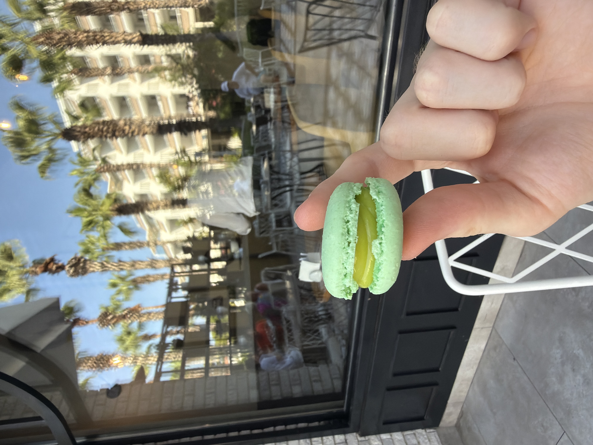 In Kemer, Turkey, a person's hand is holding a green macaron with a yellow filling in front of a cafe. The macaron is positioned in the foreground, slightly off-center. Behind the macaron, a large window reflects palm trees, a multi-story building, and the interior of the cafe. The cafe's interior shows tables and chairs, and several people are visible through the reflection, some seated and some standing. The exterior of the cafe features a black paneled wall below the window, with a light-colored tiled floor and a white chair visible in the lower right corner. The overall scene suggests a sunny day, likely in a resort or tourist area, given the presence of palm trees and the building's architecture.