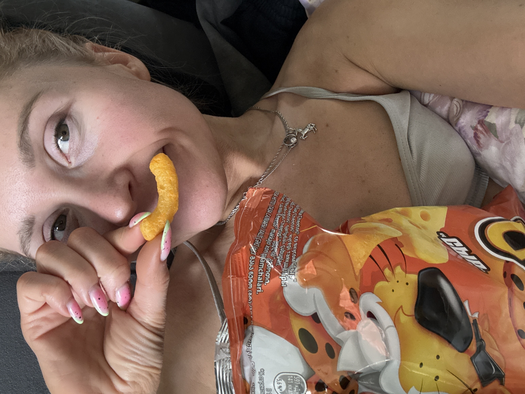 Here is a detailed description of the image:
A woman is lying down, possibly on a couch or bed, holding a Cheeto in front of her mouth. She is holding the Cheeto with her left hand, and her fingernails are painted with a pink and green watermelon design. She is wearing a thin silver necklace with a charm. Beside her is a bag of Cheetos featuring Chester Cheetah. The bag has text in various languages, including Georgian. The text indicates it is Cheetos "with Cheese flavor." The image does not contain any information that could lead to a specific location in the city of Chaikovsky, Russia.