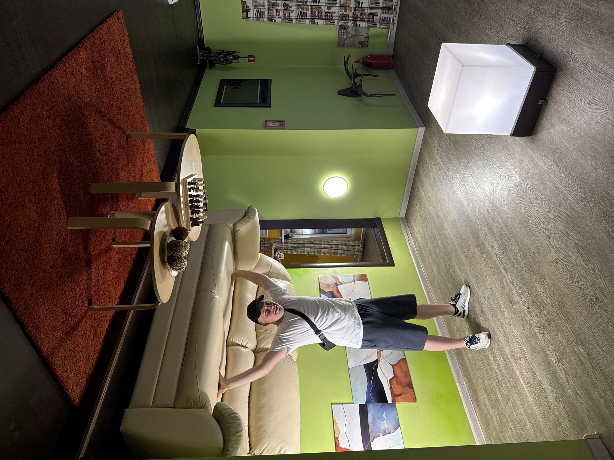 The image depicts an indoor scene within what appears to be an attraction designed to create an illusion of being upside down. A man wearing a white t-shirt, dark shorts, a black baseball cap, and athletic shoes is standing in the center of the room, holding onto the underside of a couch that is mounted to the ceiling. The couch is beige. A circular table with what looks like decorative balls and a set of chess pieces is also attached to the ceiling above the couch. The walls of the room are painted a bright green, and the floor is a wood-grain laminate. On the floor, a large, translucent white cube with a dark base acts as a light fixture. In the background, there are several abstract paintings on the wall, a fire extinguisher, and a curtain with a pattern of books. The overall impression is that the room is designed as a "house upside down" experience. The user prompt specifies the city of Kazan, Russia, but there are no visual cues within the image to confirm this location.