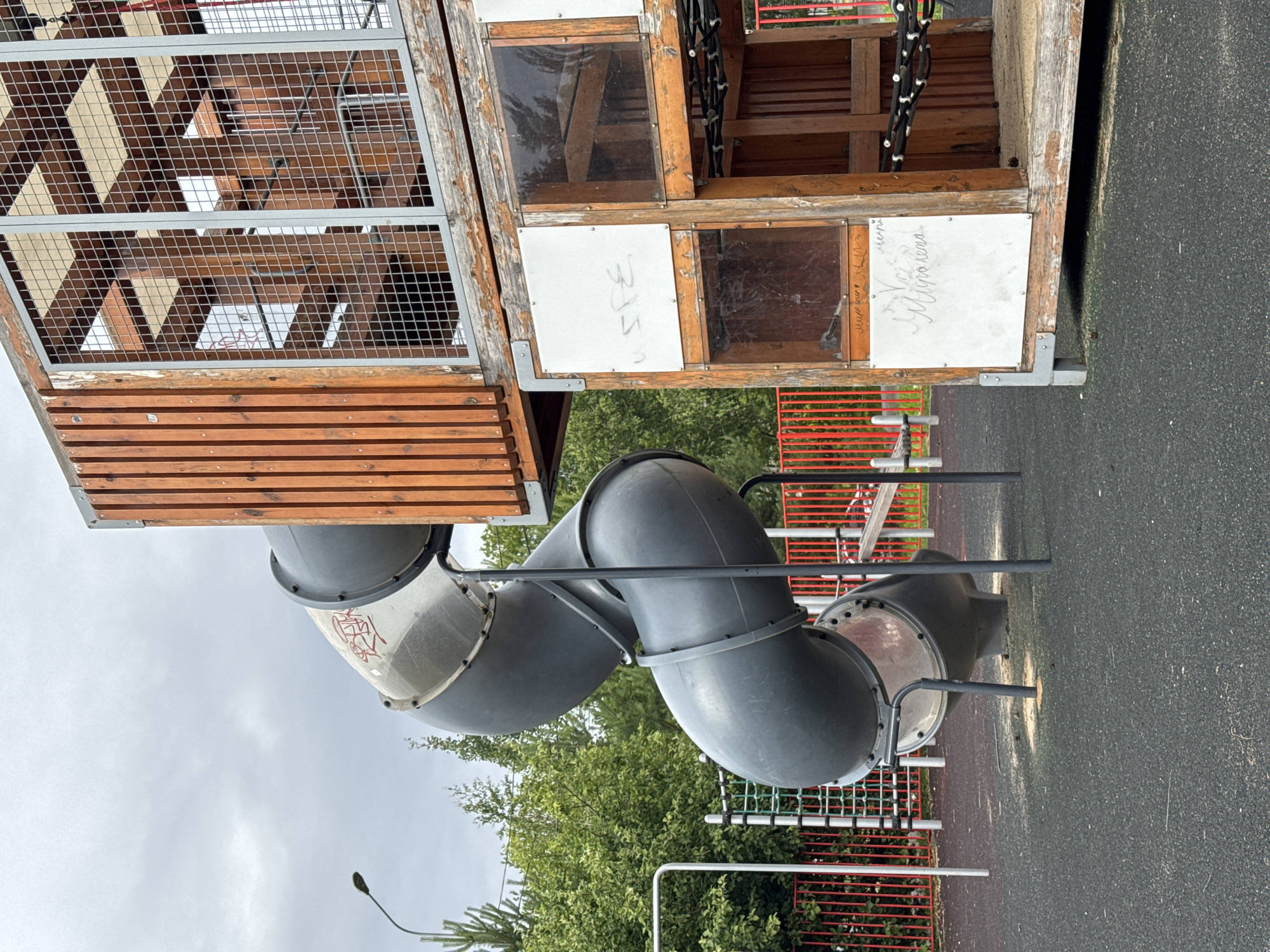 The image shows a playground structure with a wooden and metal tower that has a mesh grate on one side. Attached to the tower is a dark gray, curved slide. The slide appears to be made of plastic and has a metallic interior. There is some graffiti on the exterior of the slide. The playground is situated outdoors, with trees and a cloudy sky visible in the background. There are also red metal fences in the background, suggesting it is part of a larger park or recreational area. The ground is covered with a dark, textured surface, likely rubberized safety surfacing common in playgrounds. The overall scene suggests an overcast day. There are handwritten words visible on white panels on the wooden tower. One panel has the word "SPE" in large block letters. Another panel has cursive writing that is difficult to decipher but appears to be a name or message. A third panel also has cursive writing. The image is oriented vertically.