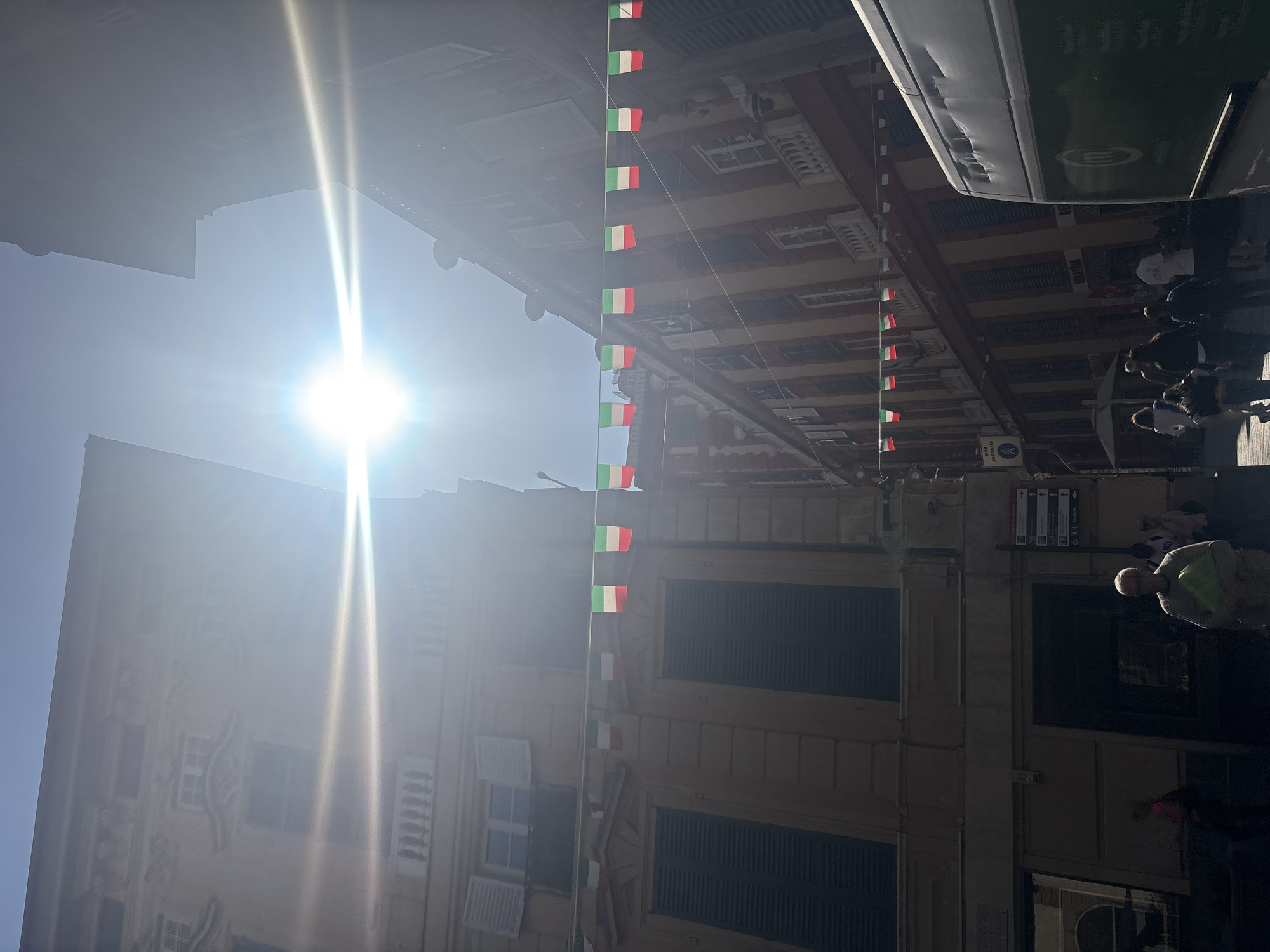 This image depicts a sunny street scene in an Italian city, likely Genoa. The photograph is taken from a low angle, looking up towards the sky and the buildings lining a narrow street.
A brilliant sunburst is prominently featured in the upper left, casting a strong glare across the sky and creating stark contrasts between intensely lit areas and deep shadows. The weather is clear and bright, indicating midday or early afternoon.
Both sides of the street are flanked by multi-story buildings with classic European architectural elements, including numerous windows, some of which have closed, louvered shutters. Extending across the street, strung between the buildings, is a long line of small Italian flags (green, white, and red bunting), adding a festive or patriotic touch to the urban environment.
Several people are visible on the right side of the street, some walking and others standing. Their figures are mostly indistinct due to shadows and distance, but one person in a light-colored top and another in a dark top are discernible. In the upper right corner, part of a large vehicle, identified as a bus, is visible. Visible text on the side of the bus includes "AMT", "VOLVO", and "IREN".
On one of the buildings to the right, a small sign features universal symbols for "Toilette" (restroom) along with directional arrows. Another sign possibly reads "via pedonale" (pedestrian street). The Italian flags and architectural style are strong indicators of an Italian location, aligning with Genoa. The overall scene suggests a vibrant, pedestrian-friendly area.