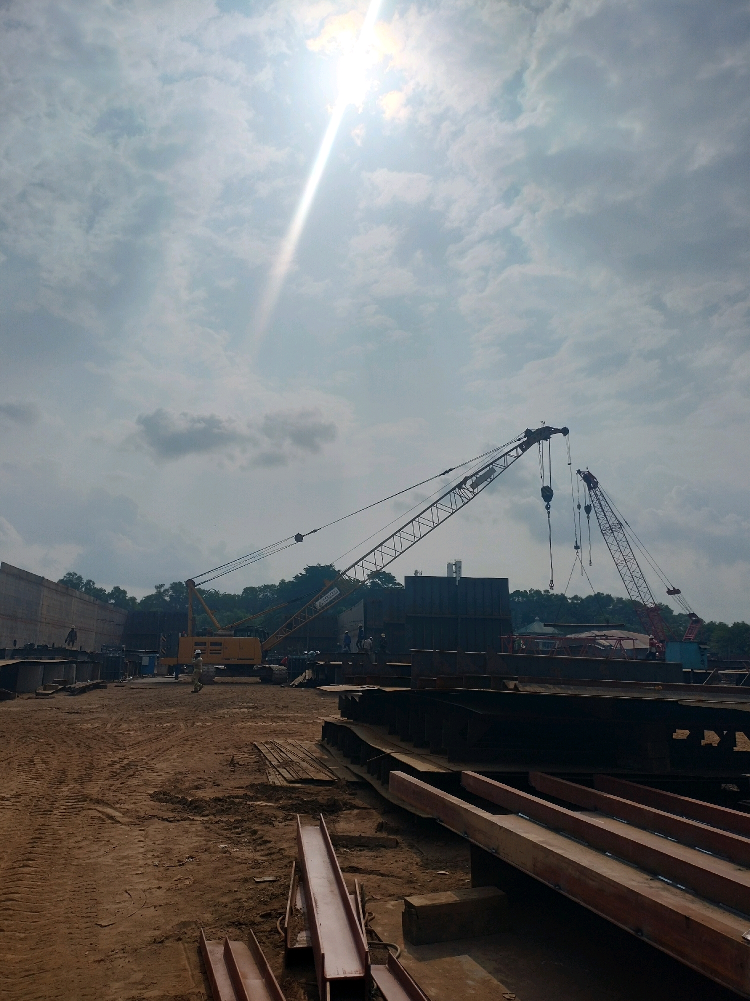 The image depicts a construction site in Batam, Indonesia, under a bright, cloudy sky with the sun prominently visible and casting rays. The foreground is dominated by a rough, dirt ground with tire tracks and scattered metallic components, likely parts of a larger structure. In the middle ground, several large cranes are positioned, with one prominent yellow crane to the left and a taller red crane to the right, both with their booms extended. The cranes appear to be in operation, with cables and hooks visible. There are also dark, rectangular structures and a concrete wall on the left side of the scene. Several individuals, dressed in work attire, are dispersed across the site, some appearing to be supervising or operating equipment. The background features a dense line of green trees, suggesting the construction site is located near a natural area. The overall scene conveys an active construction environment.