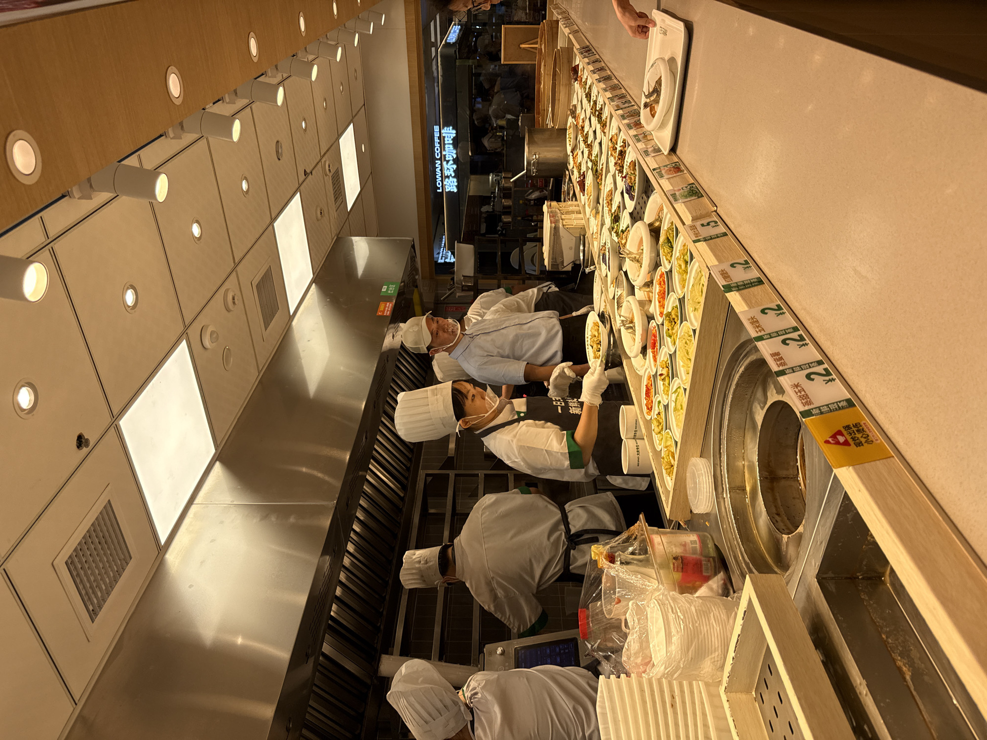 The image depicts a food service counter in what appears to be a cafeteria or a self-service restaurant in Tianhe District, China. Several individuals, dressed in chef's attire including hats and aprons, are actively working behind the counter. One person is wearing a face mask and gloves, suggesting adherence to hygiene protocols.
The counter is laden with numerous small dishes containing a variety of food items, predominantly prepared dishes. These dishes are arranged in neat rows on display shelves. The lighting in the establishment is bright, with illuminated ceiling panels and spotlights. In the background, there is a sign that reads "LOWAN COFFEE" in English and Chinese characters, indicating a coffee shop or a branded section within the establishment.
Various serving utensils and containers, such as stacks of white disposable bowls and plastic bags of food items, are visible on the counter. The overall environment suggests a busy food service operation.