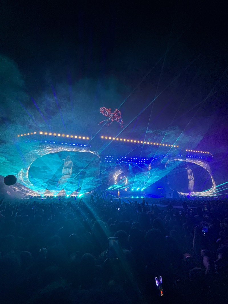 A crowd of people is gathered at what appears to be a concert in Indio, United States. A stage is set up in front of the crowd with screens displaying images of performers. Above the stage, there are lights and laser beams, and a drone display of an abstract shape.