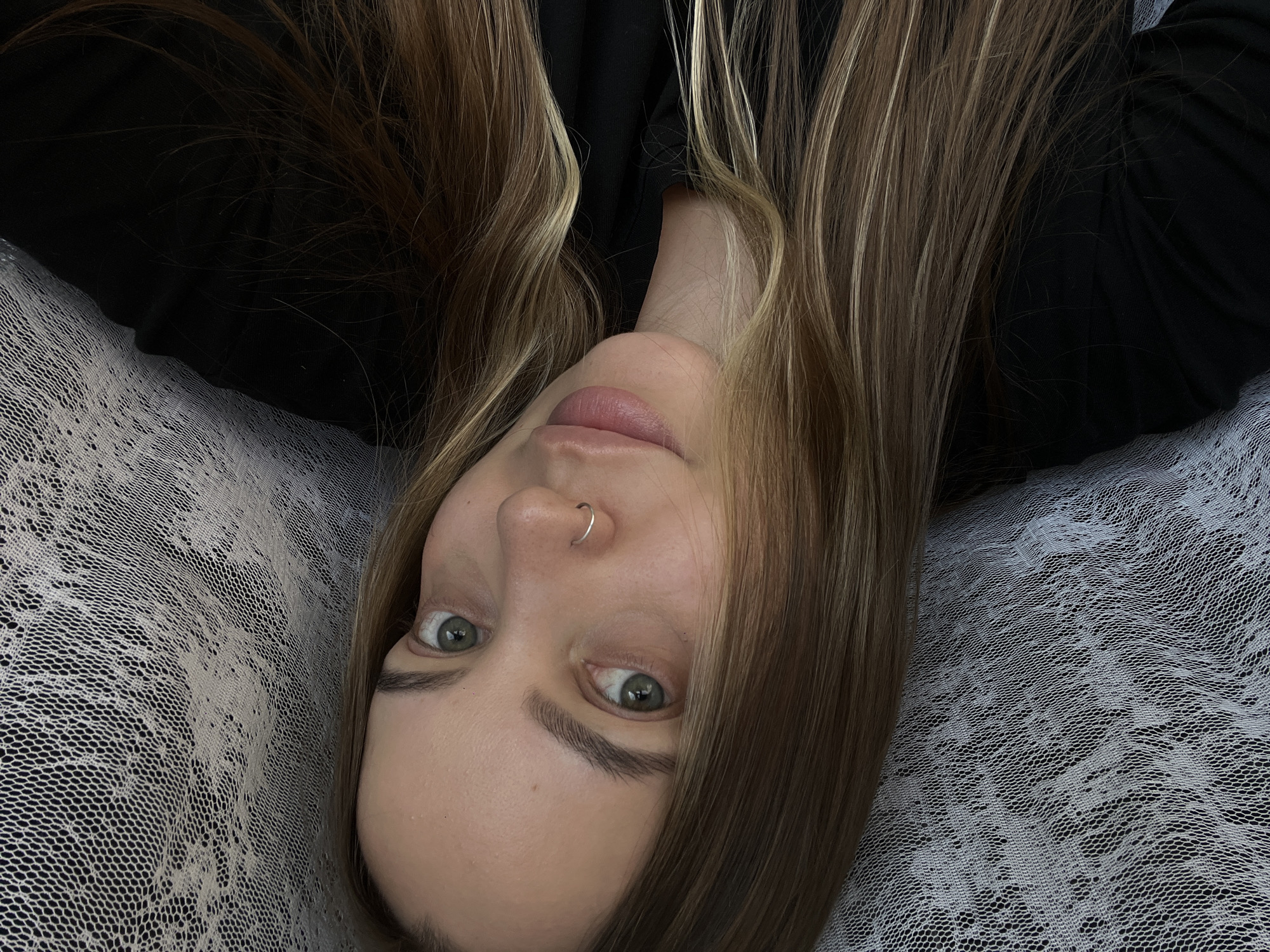 A young woman with long, blonde-brown hair and green eyes is pictured in a close-up selfie. She is wearing a black top and has a silver hoop nose ring. She is positioned in front of a white lace-like fabric that creates a textured background. The lighting is soft, with shadows accentuating her facial features. The environment appears to be indoors, possibly a bedroom or studio, given the fabric backdrop. The image does not provide any contextual information that would indicate a specific location within the city of Novopokrovskaya, Russia.