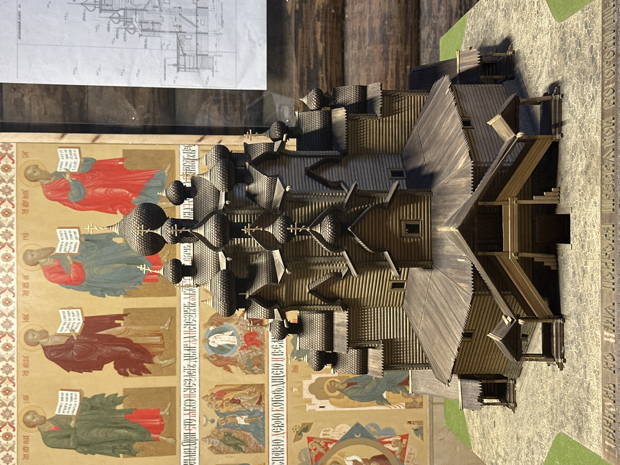 The image shows a detailed wooden model of a Russian Orthodox church with multiple onion domes and intricate architectural details. The model is placed on a textured base resembling cobblestones and grass. In the background, there are parts of what appear to be murals or religious icons depicting figures in traditional attire, and a blueprint or architectural drawing is visible on the right. The church model is the primary subject, rendered with precise craftsmanship, showcasing the tiered structure and the wooden construction. The overall scene suggests an exhibit or display, possibly in a museum or cultural center, related to Russian architecture and religious art. There are no people visible in the image.