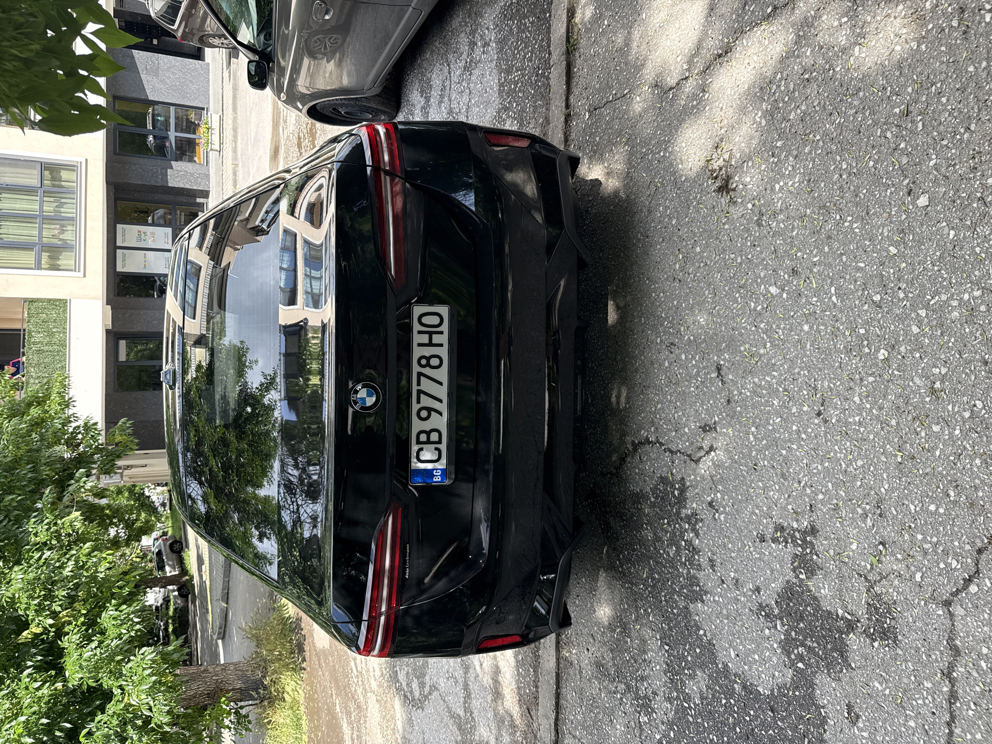 The image depicts the rear of a black BMW SUV parked on a street. The license plate reads "CB 9778 HO," indicating it is registered in Sofia, Bulgaria. The background shows buildings with reflective windows. Based on visible architectural styles and foliage, the scene appears to be set on a street in an urban area of Plovdiv, Bulgaria. The street surface shows signs of wear and tear with some darker wet patches. A portion of another vehicle, possibly a grey sedan, is visible on the upper left side of the picture. The image appears to have been taken in daytime, with shadows indicating the presence of sunlight.