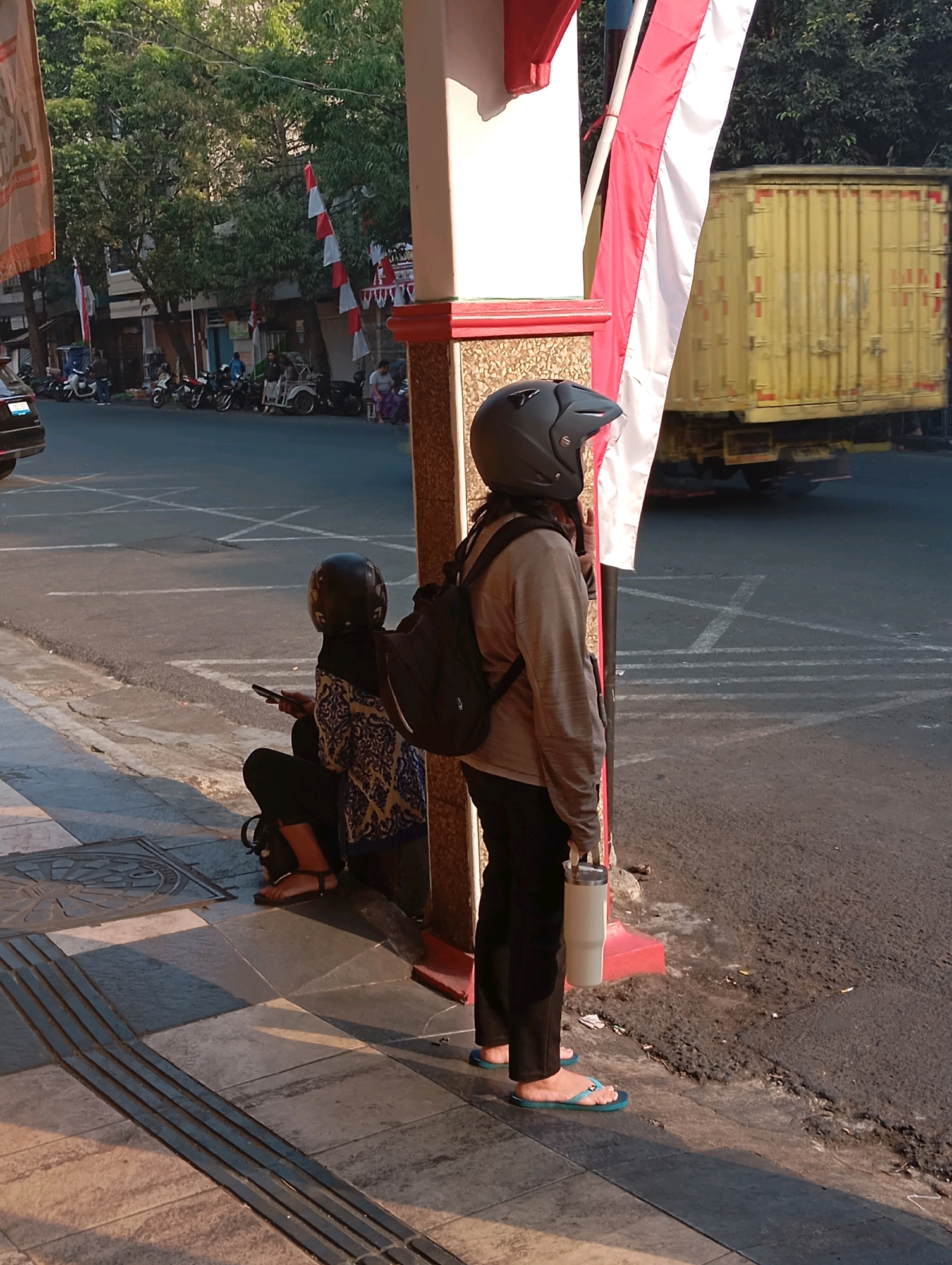 Two individuals, one wearing a helmet, are standing on a sidewalk next to a street. A yellow truck is visible in the background. The scene appears to be in the city of Malang, Indonesia, as suggested by the red and white Indonesian flags hanging in the background. The person in the helmet is holding a water bottle. The other person is seated on the sidewalk, looking at their phone. The ground is paved with stone tiles, and there are raised tactile paving strips for the visually impaired. The sun is casting long shadows, indicating it is either early morning or late afternoon.