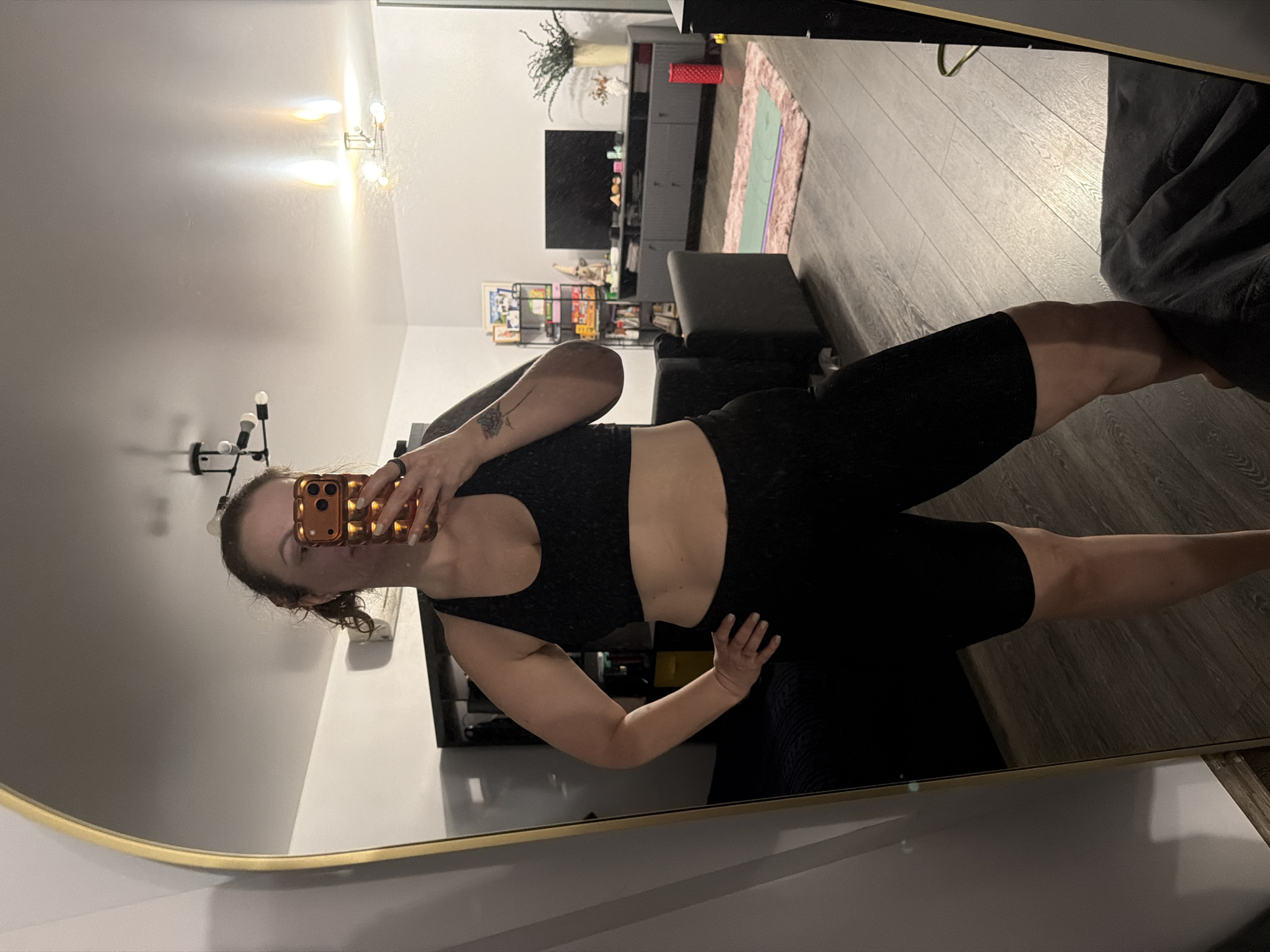 A woman is standing in front of a mirror, taking a selfie in what appears to be her living room. She is wearing a black sports bra and black shorts. The room includes a TV, shelving unit, ottoman, and a rug. The location is not identified in the city of Moscow, Russia.