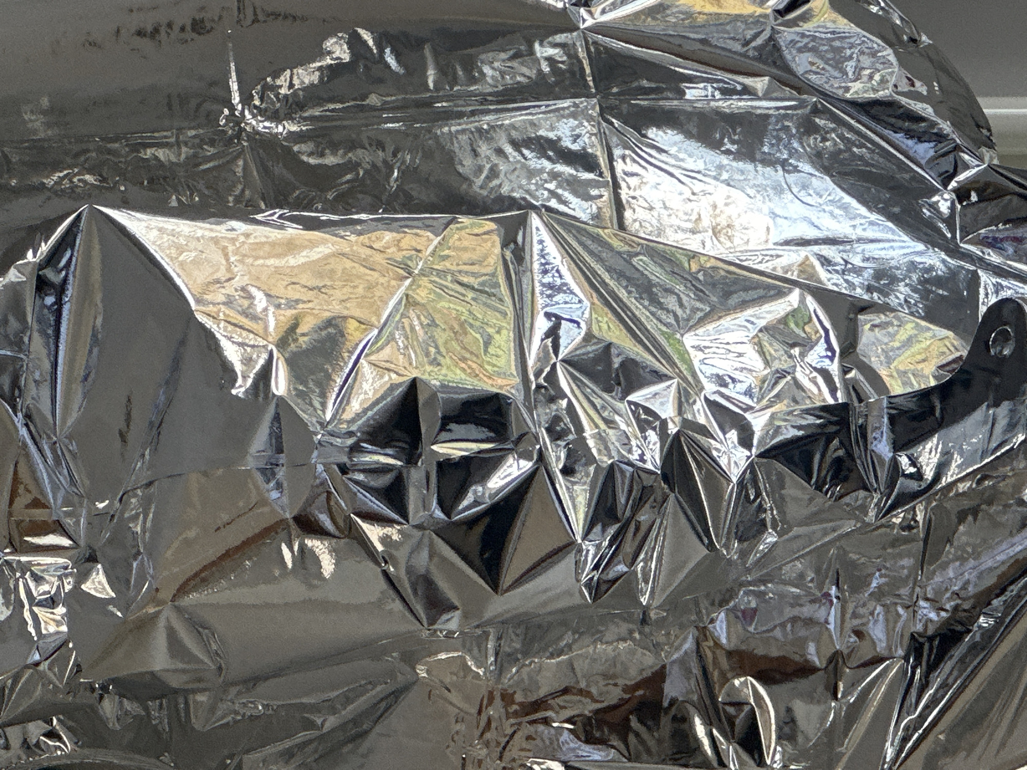 The scene, located in the city of Wealden, United Kingdom, consists of a vertical close-up of a severely crumpled, highly reflective silver material, consistent with aluminum foil or a similar metallic film. The material's surface is characterized by an intricate, chaotic network of sharp creases, deep folds, and irregular planar facets. This texture generates numerous bright highlights and dark, contrasting shadows, defining the material's three-dimensional relief. Reflections on the surface exhibit amorphous patches of bright white, yellow, and subtle green hues, indicating ambient light and potentially surrounding environmental elements, but no specific objects or figures are discernible within these reflections. There are no people, explicit objects beyond the crumpled material, actions, or interactions directly depicted within the frame.