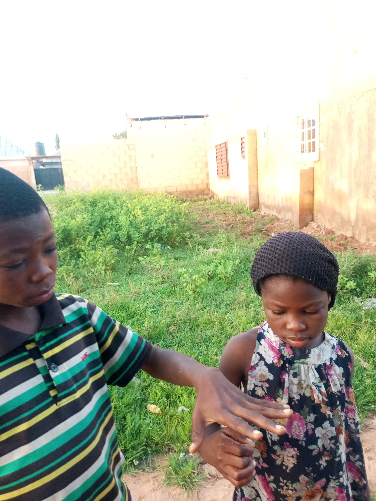 In Tempure, Nigeria, two young children are captured in an outdoor scene. A boy, wearing a horizontally striped shirt in black, green, yellow, and white, is positioned on the left. His left arm extends forward, and his hand is near the face of a girl to his right. The girl is wearing a sleeveless floral-patterned dress and a black knitted cap. Her gaze is directed downwards. Behind them, a building with light-colored walls and windows is visible, with a patch of green grass and some dry dirt in the foreground. The lighting suggests it is daytime, possibly late afternoon due to the warm tone of the light.