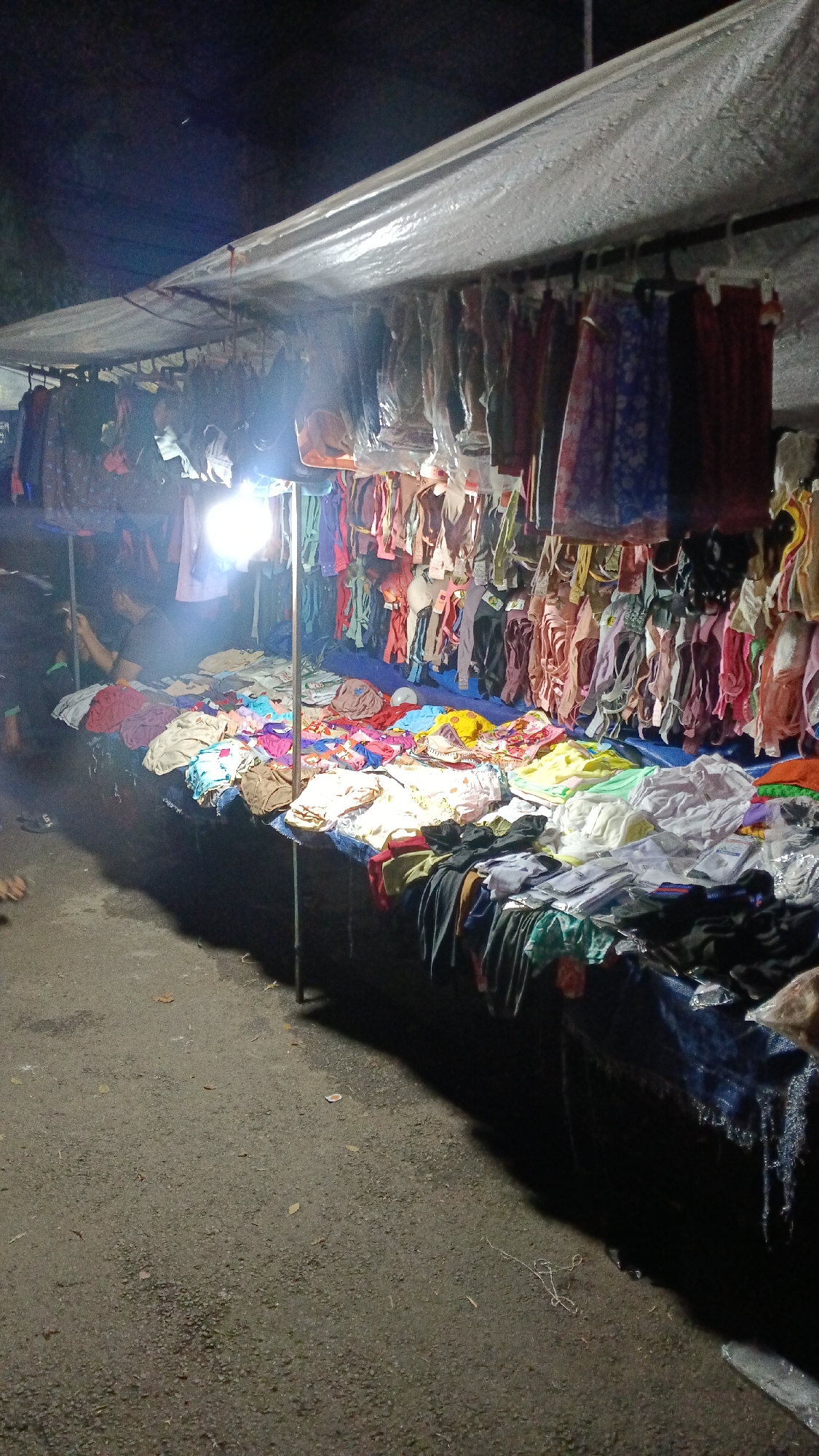 The image depicts an outdoor stall at night, likely a street market, in Tajurhalang, Indonesia. A white tarp roof is supported by metal poles, creating an overhead shelter for the displayed merchandise. The stall is illuminated by a bright, possibly portable, light source hanging from the roof.
The stall features a long, table-like surface covered in various folded clothing items. These items appear to be mostly undergarments, such as bras and panties, in a wide range of colors and patterns. Some are neatly folded, while others are more casually arranged. Behind the table, numerous pieces of clothing are hung on hangers from a horizontal bar. These hanging items include what appear to be dresses, skirts, and possibly shirts, in diverse colors and fabric textures.
In the background, a person is partially visible on the left side of the stall, seemingly engaged with their phone. Their posture suggests they might be a vendor or a shopper. The ground in front of the stall is paved with asphalt, showing some scattered leaves and debris. The overall environment is dimly lit, characteristic of a nighttime market setting, with the stall being the central focus of illumination.
