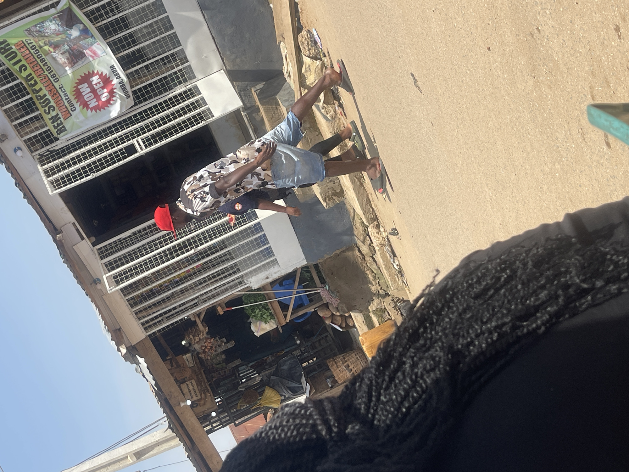 In Abuja, Nigeria, an outdoor scene unfolds with a commercial establishment in the background. A banner on the building reads "BEN SUPPER STORE WHOLESALE/RETAILER" and displays contact information "08164806072." Below this, a prominent "NOW OPEN" graphic is visible, along with "WUMBA, ABUJA." Two individuals are walking in the foreground. One person, facing away from the camera, wears a red baseball cap, a camouflage-patterned short-sleeved shirt, and denim shorts. The other person is partially visible behind them, wearing dark clothing and green slides. The ground is paved with a light-colored, sandy surface. The environment appears to be a street scene, with a building that functions as a store, suggesting a commercial area within the city.