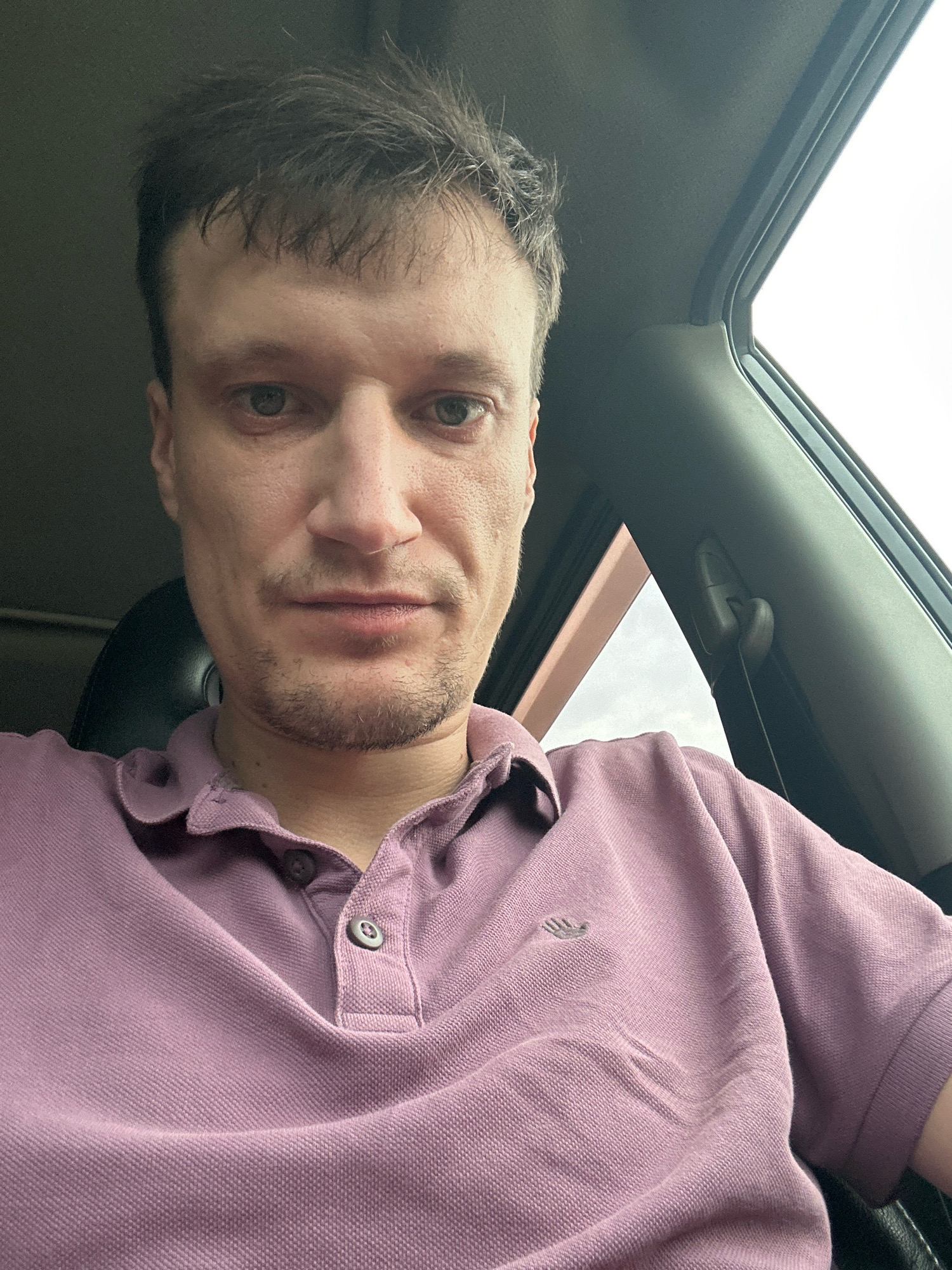 The image is a close-up, waist-up selfie of a man taken inside a car. The man is wearing a light purple polo shirt with two buttons at the collar. His hair is dark brown and messy. He has fair skin and appears to be in his late 20s or early 30s. The background shows the interior of a car, including the car's ceiling and window frame. The lighting suggests it is daytime, possibly overcast. The location of Shatava, Ukraine is not discernible from the image itself.