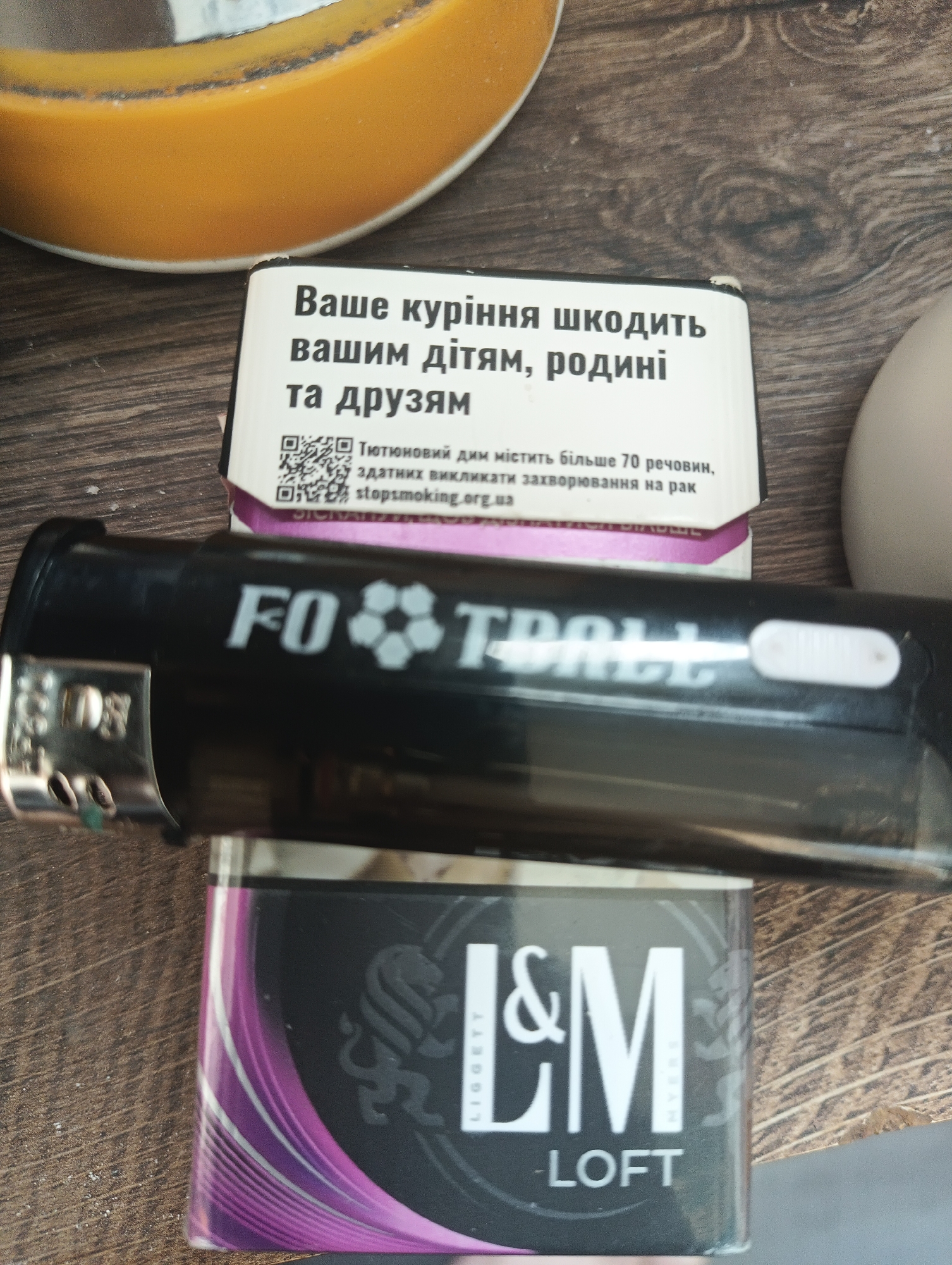 The image displays a close-up, slightly overhead view of a black cigarette lighter resting on a pack of L&M LOFT cigarettes. These items are positioned on a light brown wooden surface, likely a table.
The cigarette pack features a dark grey and purple color scheme with the brand name "L&M" and "LOFT" prominently displayed in white text. The words "LIGGETT MYERS" are partially visible in smaller print along the sides of the main logo. The top flap of the cigarette pack carries a prominent health warning in Ukrainian, which translates to: "Your smoking harms your children, family, and friends." Below this, more text states: "Tobacco smoke contains more than 70 substances that can cause cancer," followed by a QR code and the website "stopsmoking.org.ua."
The black lighter, placed horizontally across the cigarette pack, has the word "FOOTBALL" printed in white, with a stylized soccer ball icon replacing the letter 'O'. The metallic igniter mechanism of the lighter is visible on its left side.
In the upper left corner of the frame, part of a round, orange or yellow object with a darker rim is visible, potentially an ashtray or a decorative bowl. A small portion of a white or light-colored object is also seen on the right side of the wooden surface. The wooden surface itself exhibits a noticeable grain pattern.
The scene depicts an ordinary indoor setting, likely a home or a casual area, with items commonly associated with smoking. The image provides clear views of the cigarette branding and the mandatory health warnings. This scene is located in Karnobat, Bulgaria.
