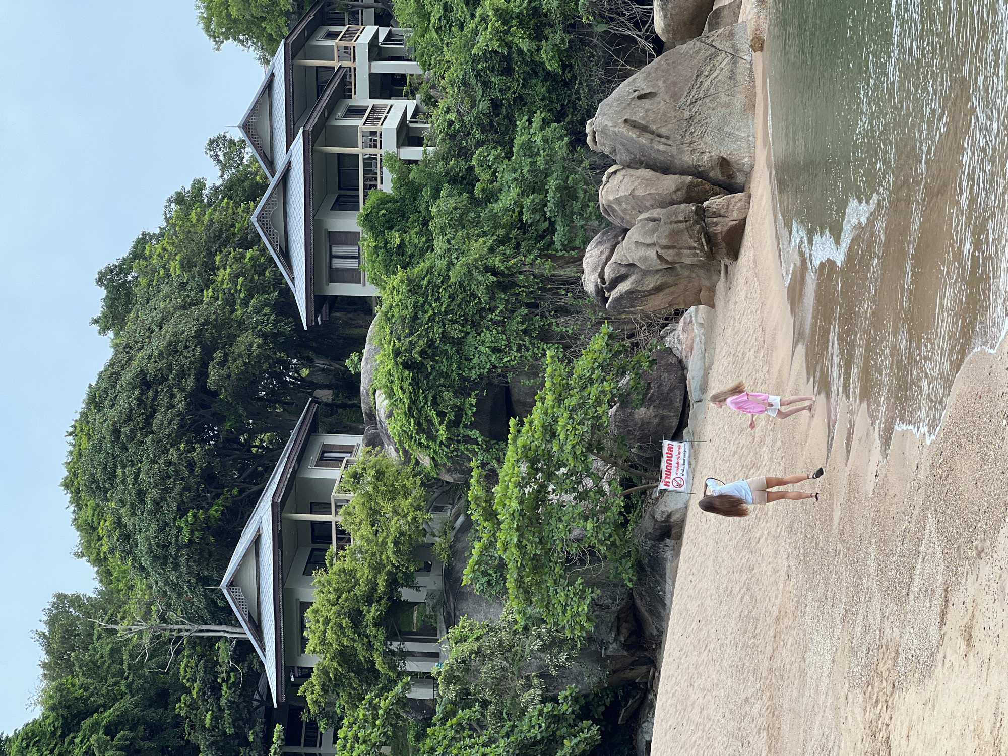 The image shows a tropical beach with two young girls on the sand near the water's edge. Behind them, several buildings are visible, built into a lush, green hillside dotted with large rocks. A sign with Thai writing is posted on the beach in front of the girls. The water is calm, and the sky is overcast. The location is Baan Chaweng Noi, Thailand.