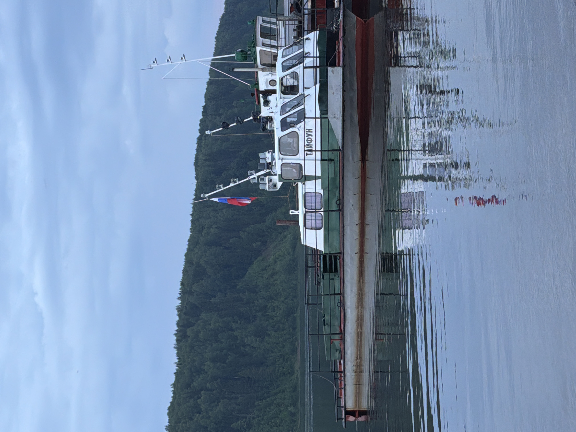 The image shows two docked boats on a body of water, likely a river, with a tree-covered hillside in the background. The boat in the foreground is green and white with a Russian flag on its mast. The word "ТАЙФУН" is visible on the side of the boat. The second boat, docked behind the first, is also white with some green accents and appears to be a similar size. The water reflects the boats and the sky. The sky is overcast. The scene is set in Tyulkino, Russia.