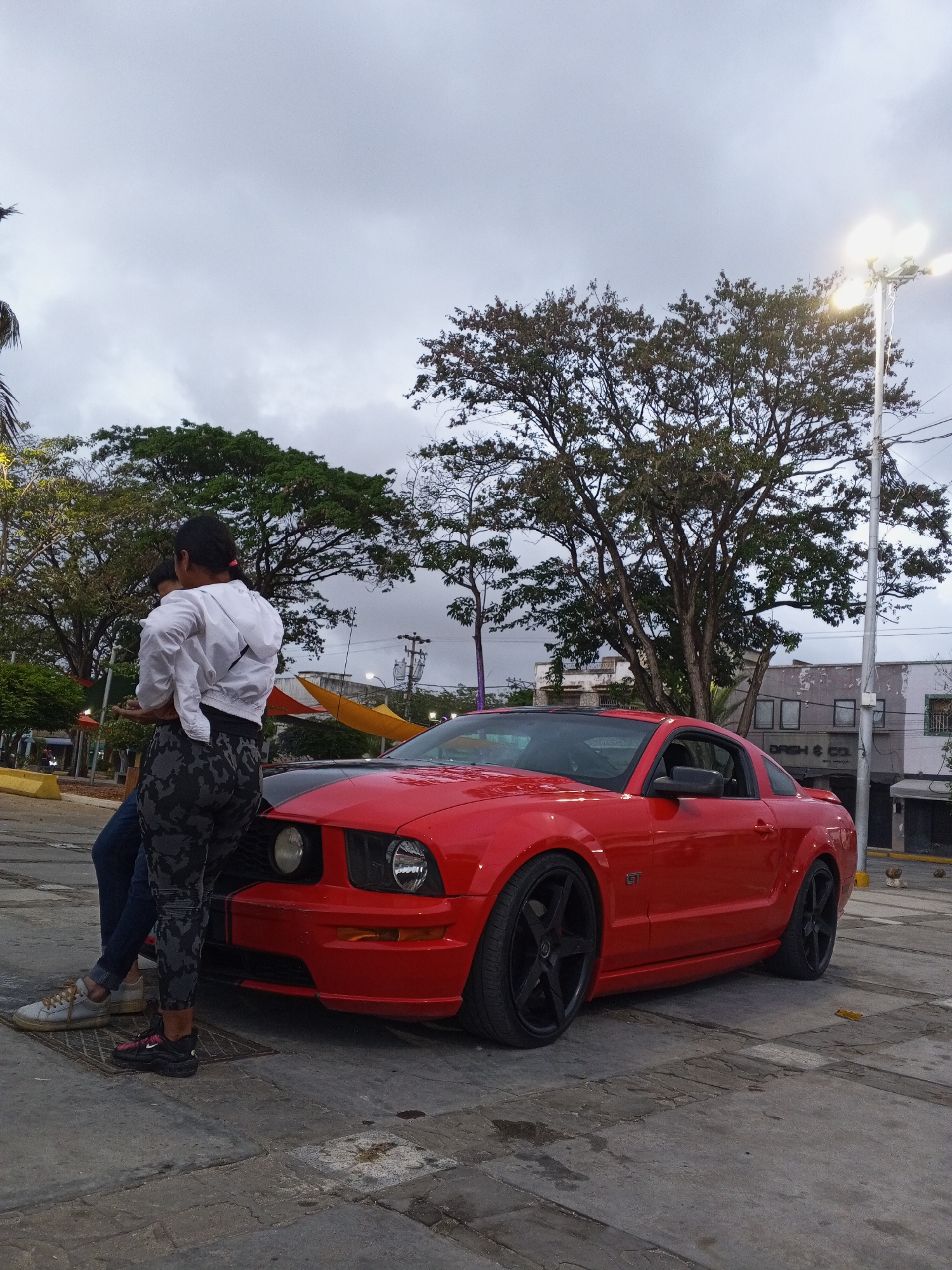 This image captures a street scene in Juan Griego, Venezuela, featuring two individuals casually leaning against a vibrant red Ford Mustang GT. The person closer to the viewer is seen from behind, wearing a white hooded jacket, dark top, and grey camouflage-patterned leggings with black athletic shoes. Another individual, partially obscured, is seated or leaning against the car's front fender, wearing blue jeans and light-colored sneakers.
The car, a red Ford Mustang GT, features a black racing stripe running over its hood and roof, and is fitted with distinctive black multi-spoke rims. A "GT" emblem is visible on the side panel.
The setting is an outdoor urban space, possibly a town square or roadside area, with worn concrete pavement. Several large trees with green and sparse foliage frame the scene. The sky is overcast and grey, indicating a cloudy day or perhaps dusk, as an illuminated street light with multiple bright lamps is visible in the background. Distant buildings, some appearing somewhat dilapidated, line the background. Text "DASH & CO." is discernible on one of these structures. The overall mood is relaxed and informal.