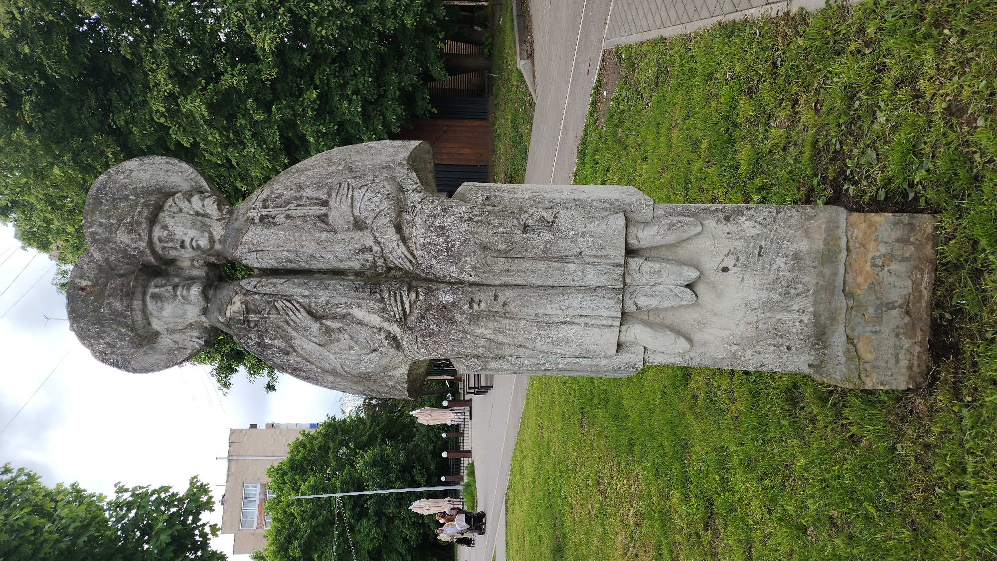 A vertical outdoor shot shows a weathered gray monument of two figures standing side-by-side on a plinth. The figures appear to be dressed in robes and hats, their hands clasped in front of them. A pathway, lined with green grass, is visible in the background. Beyond the pathway, trees and buildings can be seen. In the distance, white umbrellas suggest a cafe or outdoor seating area. The image is likely from Smolensk, Russia, given the monument style and architectural elements. There is no interaction or specific actions visible except for people sitting by the outdoor umbrellas.