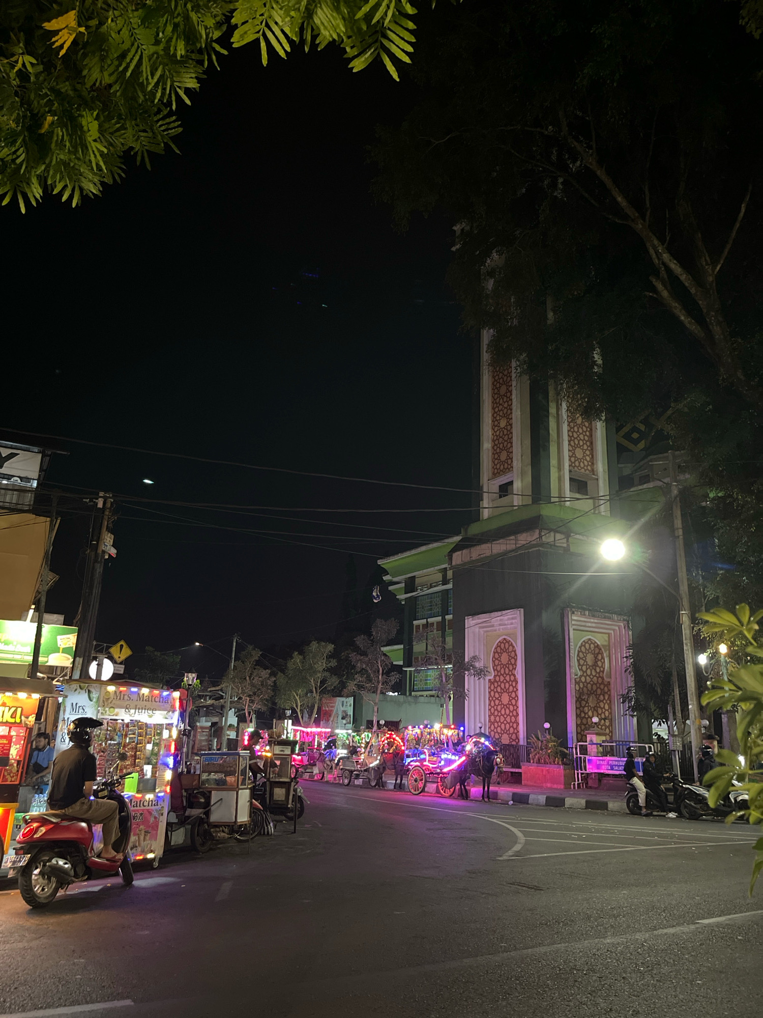 This is a night-time exterior street scene in Salatiga, Indonesia, focusing on a busy intersection. In the foreground, a red scooter is parked facing left, with a person wearing a dark shirt and light-colored pants seated on it, facing away from the camera. To the left of the scooter is a brightly lit food cart displaying various products and signage, including "Mrs. Matcha & Juice." Behind the scooter and food cart, the street continues with more food carts and several horse-drawn carriages decorated with colorful lights, suggesting a festive atmosphere. The carriages appear to be stationary or moving slowly. In the background, a tall, ornate building, likely a mosque given its architectural style and the location, is visible. The building features prominent minaret-like structures with intricate patterns and is illuminated. Trees with dark foliage are present on the right side of the frame, partially obscuring the view of the building. Power lines are strung across the scene, and streetlights provide additional illumination. The road surface is asphalt, and there are painted lane markings. People can be seen standing near the food carts and sitting on motorcycles. The overall impression is that of a lively night market or a popular gathering spot in the city.