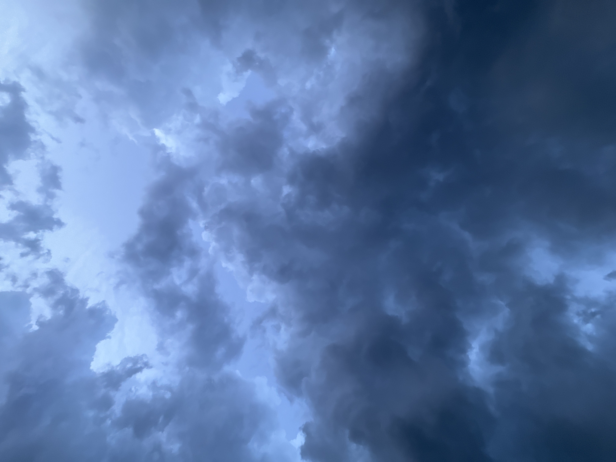 The image depicts a dramatic sky filled with dark, ominous storm clouds. The clouds are dense and turbulent, with varying shades of gray and deep blue, suggesting an impending storm. Patches of lighter sky peek through, creating a sense of depth and contrast. The perspective is looking upwards, emphasizing the immensity and power of the clouds. There are no people or objects visible, making the sky the sole subject. The overall mood is one of intensity and anticipation, likely evoked by the approaching weather. The image captures a moment of natural drama, with the clouds swirling and gathering in a powerful display. The lighting suggests it could be late afternoon or early evening, or simply a very overcast day. While no specific location cues are visible within the frame itself, the provided context places the scene in Baan Chaweng Noi, Thailand. The weather appears stormy or about to become stormy.