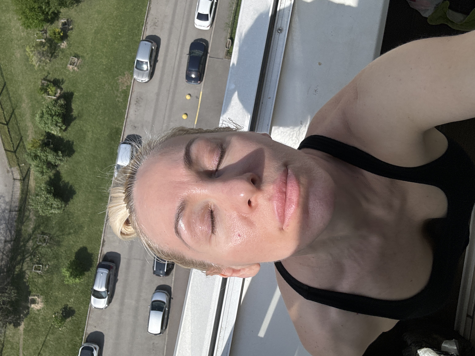 A woman is captured from a high angle, seemingly taking a selfie or recording a video. She is positioned on a ledge or balcony overlooking a street with cars parked alongside a patch of greenery. The woman has fair skin, blonde hair pulled back into a bun, and is wearing a black tank top. Her eyes are closed and her face is directed toward the camera. Below her, a street showcases parked cars, with a green area containing trees and shrubs running parallel to it. The specific location is not provided in the image.