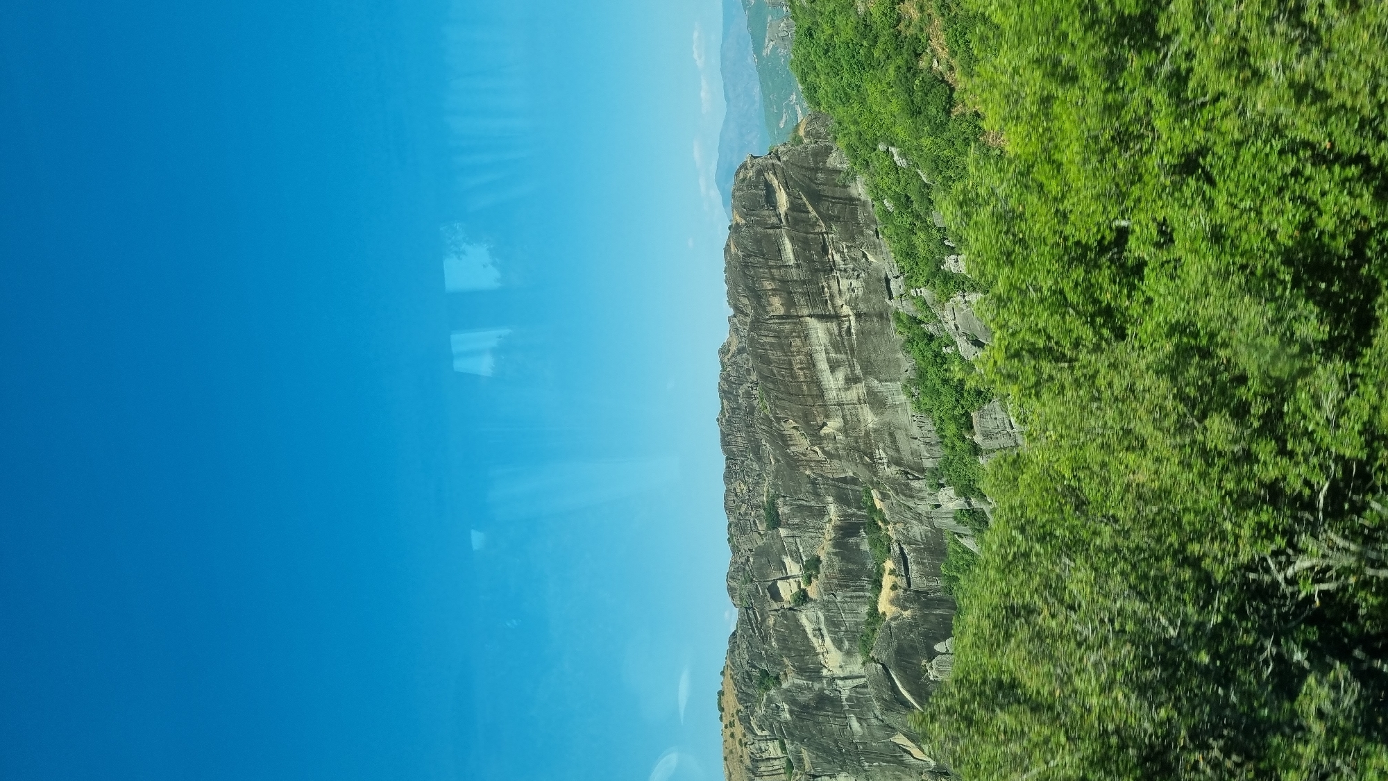 The image depicts the unique geological formations of Meteora, located near the city of Kalambaka, Greece. The foreground is dominated by lush green trees, suggesting a natural, possibly rural or mountainous, environment. Behind the trees, the towering rock pillars, characteristic of Meteora, rise dramatically. These are massive sandstone cliffs, weathered and eroded over time, with a layered appearance and a greyish-tan color. Some vegetation, appearing as small bushes and dry grass, can be seen growing on the slopes and ledges of these rock formations. In the distance, rolling hills and mountains fade into a hazy blue, under a clear, bright blue sky. The sky appears slightly distorted, possibly due to reflections on a window. No people or structures are visible in this particular frame.