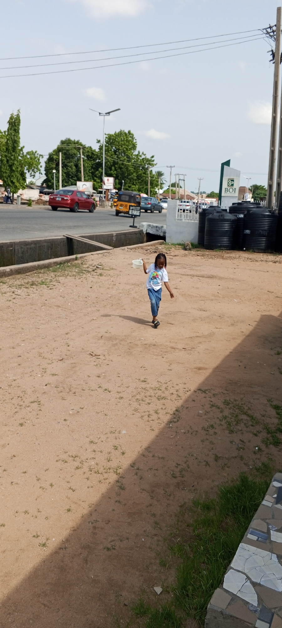 In Jalingo, Nigeria, a young Black child with braided hair, wearing a white t-shirt with graphics and blue jeans, is captured walking across a dirt path. The child is holding a white plastic bowl in their right hand and is leaning forward, suggesting they are in motion, possibly running. The environment features a road in the background with several vehicles, including a red car, a yellow auto-rickshaw, and a silver sedan. A traffic light is visible on the road. Adjacent to the road are buildings and trees, and a sign that reads "NO PARKING." In the foreground, large black water tanks are stacked, and a concrete drainage channel runs alongside the path. The ground is covered in light brown dirt with sparse patches of green grass. The scene is illuminated by daylight, casting a shadow of the child on the ground.