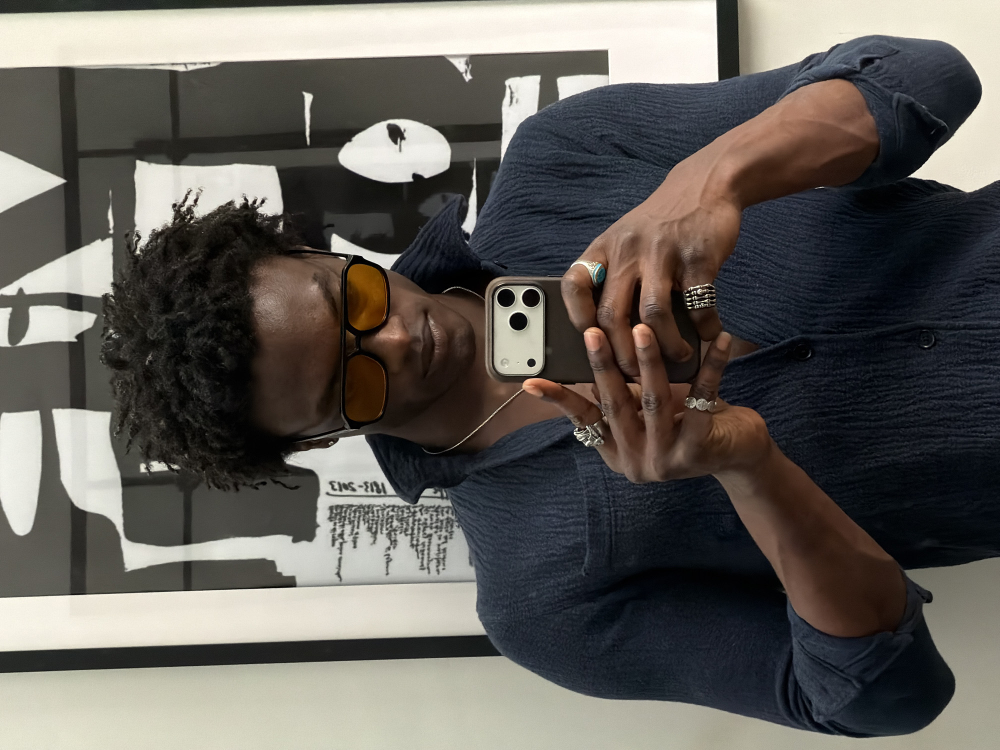 A person with dark skin, wearing a dark blue textured shirt and yellow-tinted rectangular sunglasses, is taking a mirror selfie with a smartphone. The image is captured from a slightly high angle, showing the person holding the phone with both hands, angled towards their face. Their dark, short, curly hair is visible.
The person is adorned with several rings: on their left hand, a ring with a turquoise-like stone on the ring finger and a stack of rings on the middle finger. On their right hand, a distinctive, possibly skeletal or geometric ring is on the ring finger, and another simple band is on the pinky. A thin silver chain necklace is also visible around their neck. The sleeves of their shirt are rolled up, revealing muscular forearms with prominent veins.
The background features a large, framed abstract artwork in black and white, composed of bold geometric and organic shapes. Some inverted text is visible on the artwork: "EINS-EINS" and "1813-5/13," along with other smaller, illegible text. The setting appears to be indoors, with bright, even lighting, suggesting a well-lit interior space. No specific location cues beyond the artwork are visible to confirm the London context.