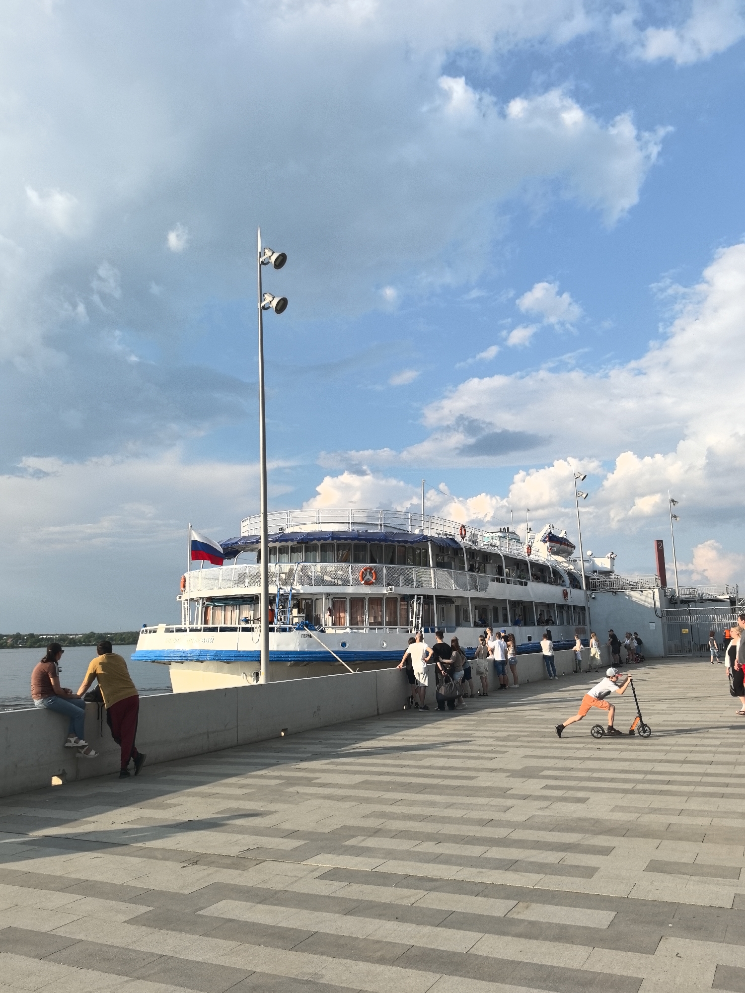The image shows a sunny day at a waterfront location in Perm, Russia, with a riverboat moored alongside a gray paved pedestrian area. The riverboat, "Пермь", has a Russian flag displayed. On the waterfront, several people are present; two individuals are sitting on the edge of a raised concrete barrier, while others are standing near the boat. A person is riding a scooter on the paved surface. Light poles are positioned along the waterfront. The sky is partly cloudy.