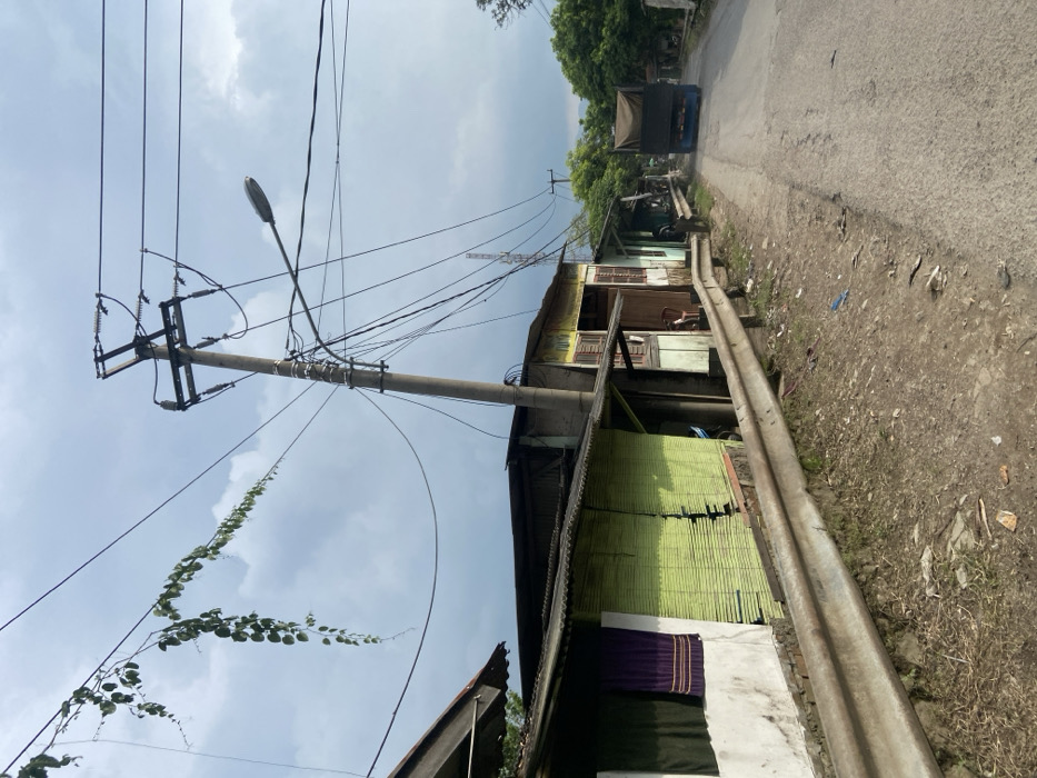 The image shows a street in Margaasih, Indonesia (latitude: -6.943, longitude: 107.556). The street is lined with small, simple houses with corrugated metal roofs. A large truck is driving down the street. The street is paved with asphalt, and the surrounding terrain is dirt. A utility pole with many power lines runs down the left side of the street. A streetlight is attached to the pole. A metal guardrail separates the street from the houses. There are several plants, vines and trees growing on the houses and the ground. The sky is cloudy.