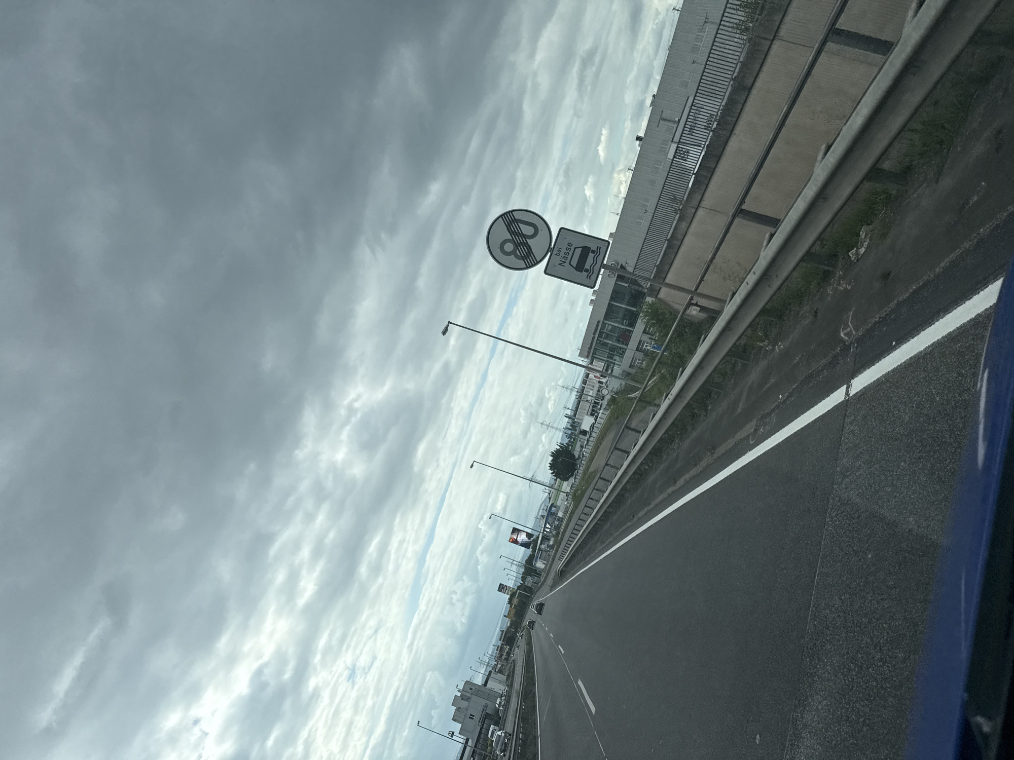 The image captures a view from inside a vehicle driving on a multi-lane highway in Koblenz, Germany. The sky is overcast with thick, gray clouds. On the right side of the highway, there are two traffic signs mounted on a pole. The upper sign is circular with a red border and a black horizontal bar, indicating an end to a speed restriction. The number "80" is superimposed on the bar. The lower sign is square and depicts a car driving through a puddle, with the German word "Nässe" (wetness) below it. This sign signifies a speed limit of 80 km/h when the road is wet. A metal guardrail runs along the side of the highway, separating it from a concrete barrier and some vegetation. Beyond the barrier, there are buildings, including what appears to be a commercial establishment with a large glass facade, and several tall light poles. Cars are visible on the highway ahead, traveling in the same direction as the viewer's vehicle. The road surface appears to be asphalt, with visible lane markings. The overall impression is a typical highway scene on a cloudy day.