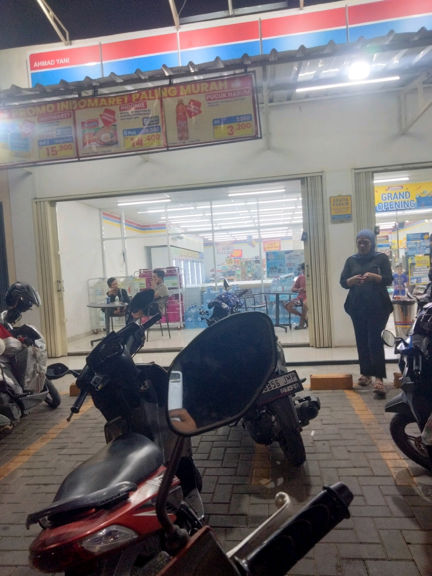 In Suka Sari, Indonesia, at night, a street-level view captures the facade of an Indomaret convenience store. The store's exterior is illuminated by artificial lights, highlighting promotional banners. One banner reads "PROMO INDOMARET PALING MURAH" (Indomaret's Cheapest Promo) and displays discounted items like Indomie noodles and bottled drinks with prices such as 15,900, 74,400, and 3,200. To the right of the main entrance, a "GRATIS PARKIR" (Free Parking) sign is visible, and further to the right, a large banner announces "GRAND OPENING." The store's large glass doors and windows reveal the interior, where bright fluorescent lights illuminate shelves stocked with products and several large blue water bottles are stacked.
Outside the store, several motorcycles are parked on a paved area marked with yellow lines. Two men are seated at a small round table near the entrance, appearing to be engaged in conversation or taking a break. A woman wearing a hijab and dark clothing stands in front of the store, looking towards the camera. The foreground is dominated by the partial view of a red and black motorcycle. The overall scene suggests a typical commercial street environment at dusk or night.