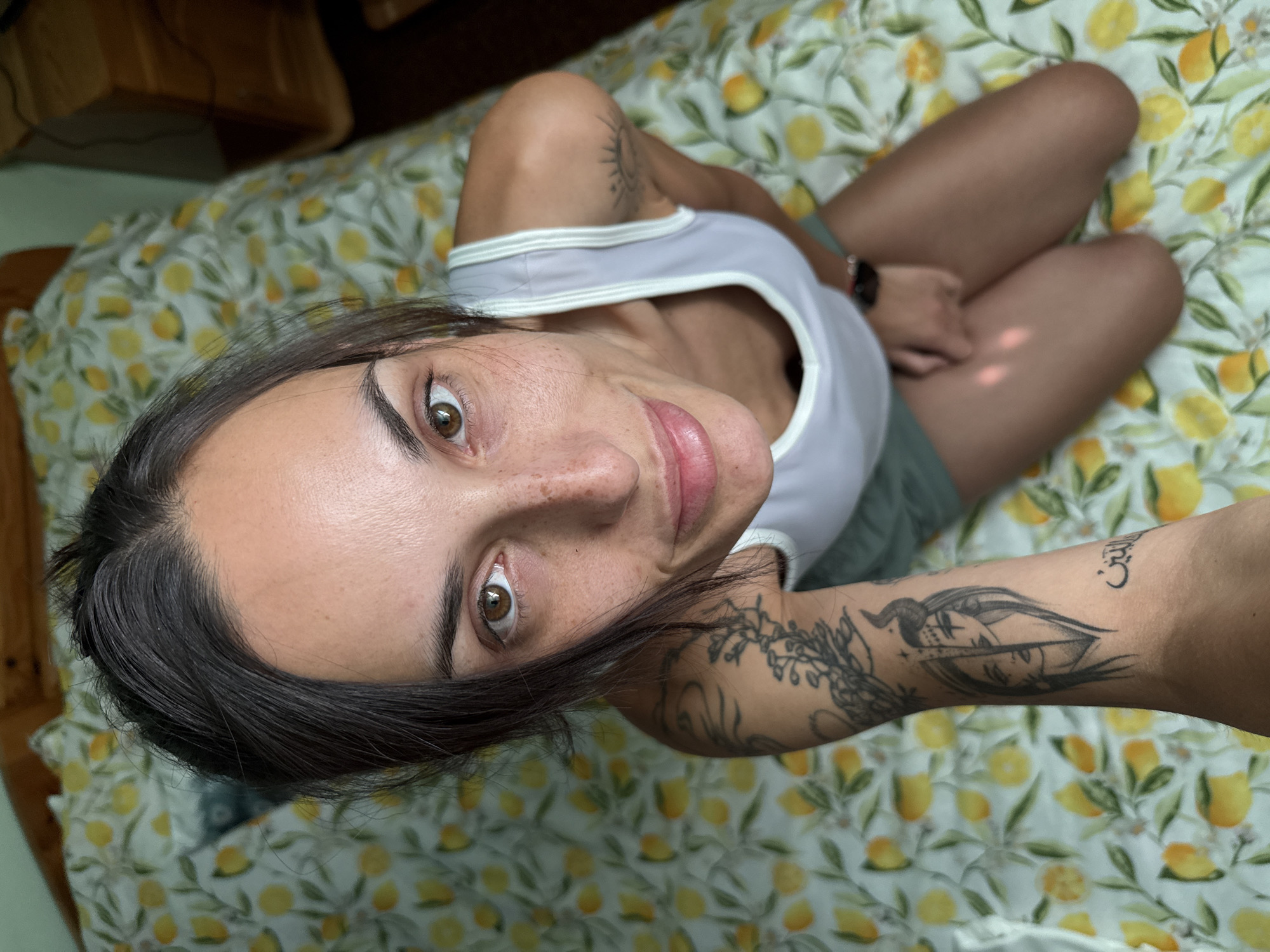 A light-skinned woman with brown hair and eyes is centered in the frame, positioned on a bed with lemon-patterned sheets. She wears a grey tank top and is seated with her knees bent. Visible tattoos adorn both arms. The background includes a partial view of a wooden structure in the upper left corner. No information indicates a location in Pleven, Bulgaria.