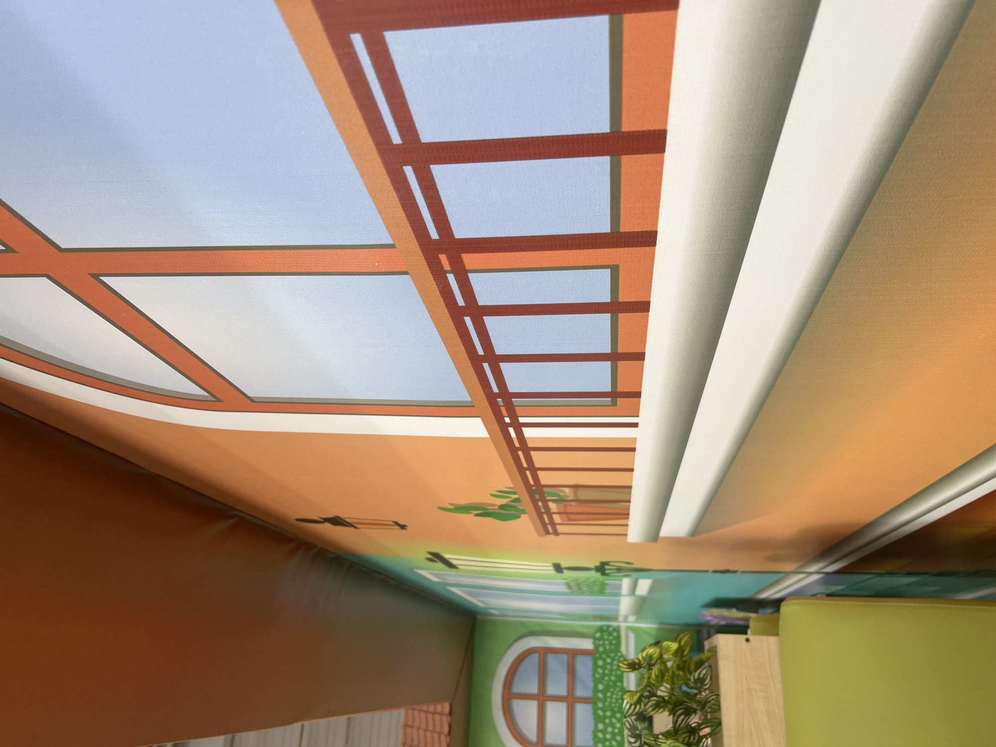 The image captures an indoor scene in Chaikovsky, Russia, featuring a large-scale, colorful mural on a wall, designed to resemble an architectural streetscape. The mural depicts a dominant orange building facade on the right, showcasing a large arched window with a reddish-brown frame and a light blue background simulating the sky. Below the window, a reddish-brown balcony railing is rendered above a white, projecting balcony ledge. Further into the painted perspective, a green building facade with a smaller arched window and depicted green shrubbery in a planter box is visible on the left. The mural also includes details such as illustrated streetlamps and potted plants on the building surfaces. In the immediate foreground, a real, lime-green upholstered seating unit is partially visible at the bottom left. Above this seating, a light wooden planter box holds a living plant with variegated green and yellow foliage, providing a three-dimensional element against the two-dimensional artwork. A flat, orange-brown ceiling or bulkhead extends across the top of the frame. The image is captured from an angled perspective, looking along the length of the mural, creating an illusion of depth within the indoor space.