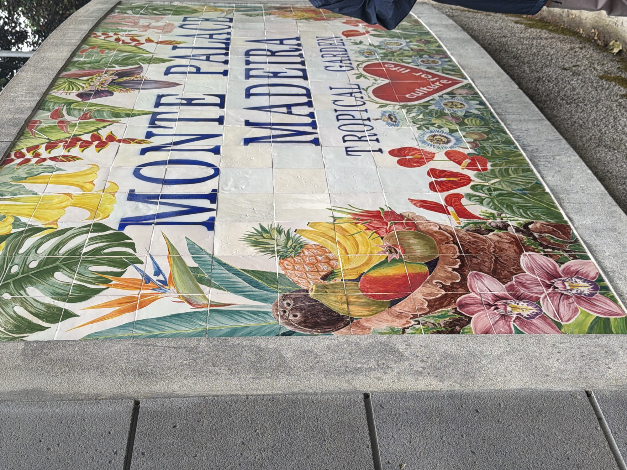 The image shows a tiled mural on a low platform in Funchal, Portugal. The mural depicts the words "MONTE PALACE MADEIRA TROPICAL GARDEN" in dark blue block letters, arranged vertically. The background of the mural is filled with colorful illustrations of tropical plants and fruits, including monstera leaves, yellow trumpet flowers, bird-of-paradise flowers, pineapples, bananas, mangoes, and orchids. A portion of a dark blue garment is visible in the upper right corner of the image. The platform surrounding the mural is made of gray concrete.