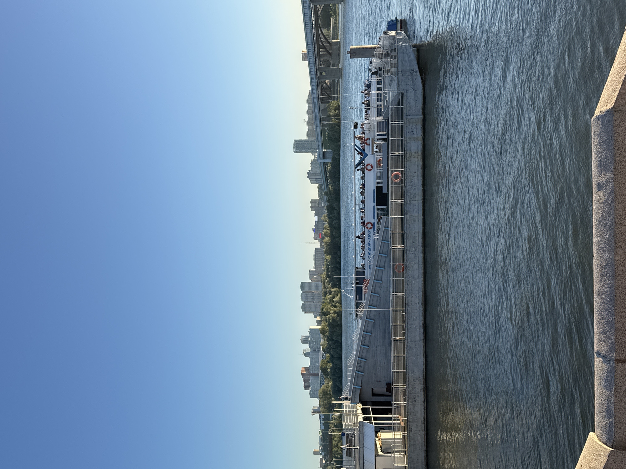 This media file depicts a clear, sunny day on a body of water, likely a river, in Novosibirsk, Russia. The scene is dominated by a long, narrow boat or barge with multiple levels, carrying numerous passengers. The passengers appear to be standing and observing the surroundings, suggesting a leisurely activity such as a sightseeing tour.
The boat's structure is industrial and functional, with a concrete-like exterior and metal railings. Several life preservers are visible along the sides of the vessel, indicating safety measures. The water is a deep blue-green and appears somewhat choppy, reflecting the daylight. In the background, a cityscape of high-rise buildings and trees lines the riverbank, with a prominent bridge arching over the water in the distance. The time of day is evident from the bright sunlight and clear blue sky, suggesting it is daytime, possibly late morning or afternoon.
On the side of the boat, the text "SKBA-193" is visible. The overall mood is calm and observational, with people engaged in taking in the sights of the city and the river. The presence of the bridge and the distinct urban skyline firmly place the location in Novosibirsk. The right side of the image shows a stone structure, possibly a quay or embankment, adding to the urban waterfront context.