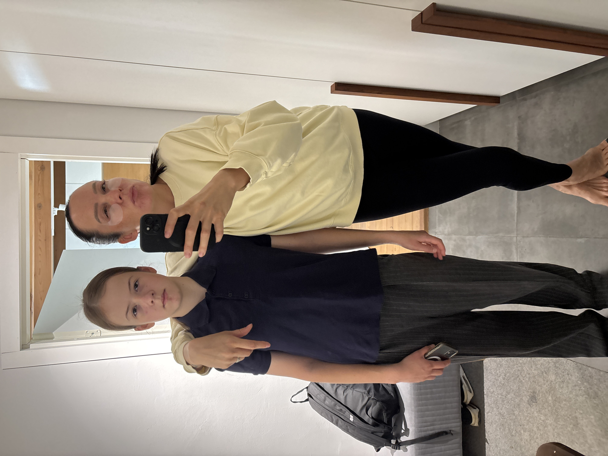A mirror selfie shows an adult woman with a light yellow sweatshirt and black leggings, wearing an under-eye patch on her left eye, holding a black smartphone and hugging a young person with light skin. The young person is wearing a dark blue polo shirt and grey pinstripe trousers. A black backpack lies on a grey ottoman or couch. The scene is indoors and looks like a residential hallway. The location is Chaikovsky, Russia.