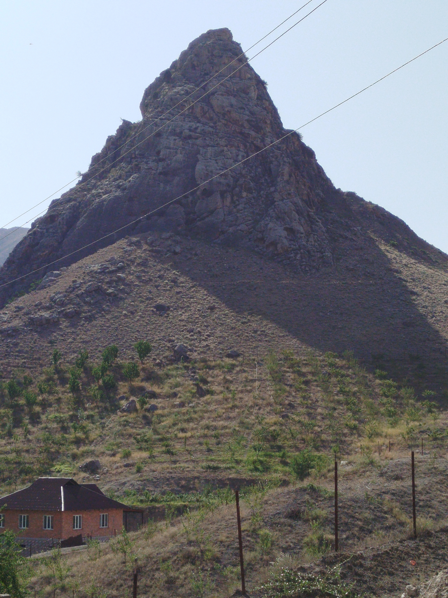 A steep, rocky mountain dominates the background of this outdoor, daytime scene. The mountain is dark gray and has a jagged peak. Several power lines stretch across the sky in front of the mountain. The foreground is a brown, dry, grassy hillside with scattered rocks and small trees. In the lower left corner of the image, there is a single-story, brick house with a dark-colored roof and several white-framed windows. The house is positioned on the hillside. The overall impression is a rugged, somewhat arid landscape with a solitary dwelling.