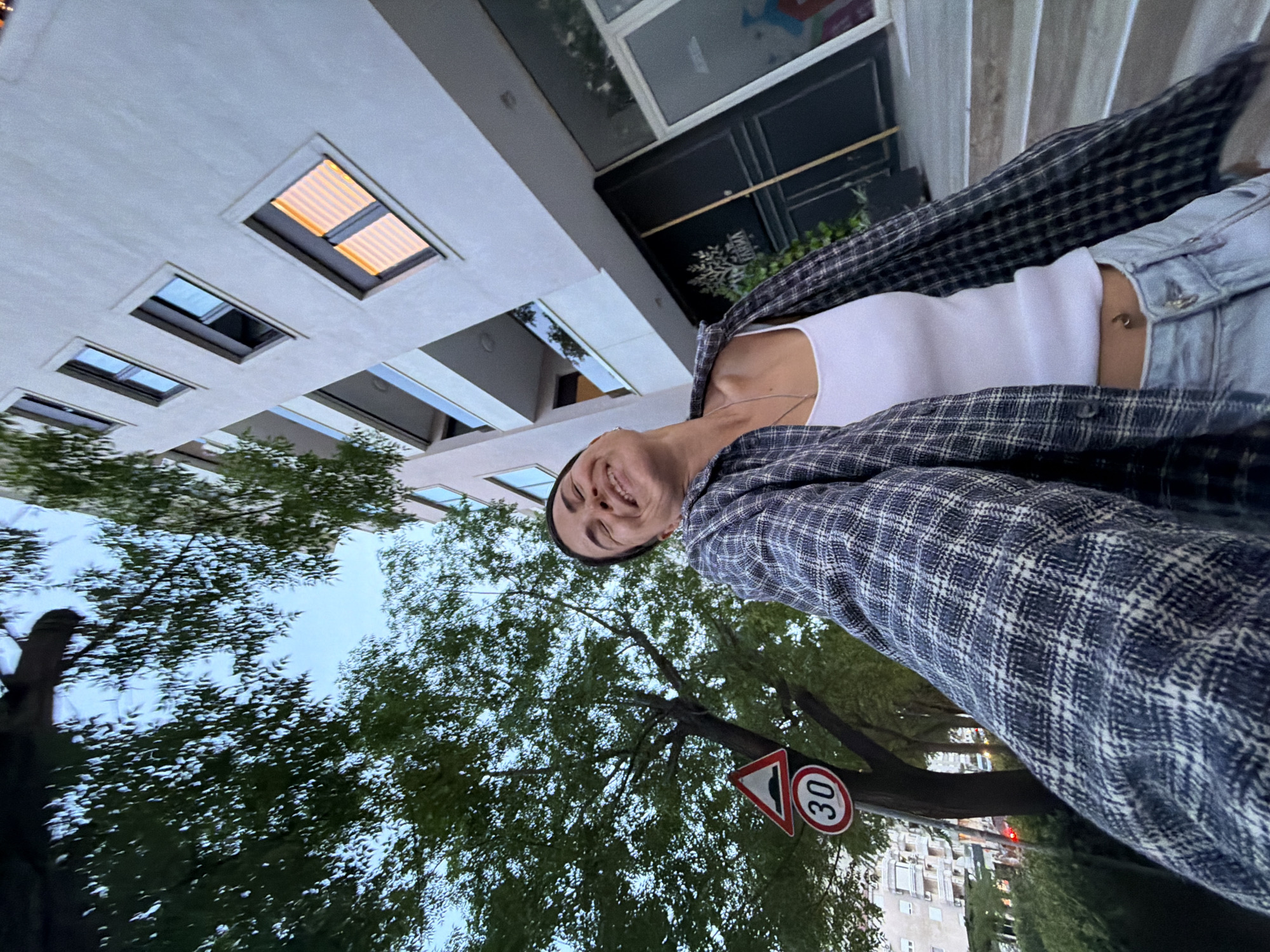 A rotated selfie captures a young woman smiling broadly outdoors in an urban setting, likely during the late afternoon or early evening in Budva, Montenegro. She looks directly at the camera with her eyes closed in a wide, joyful smile, conveying a happy and relaxed mood. She is wearing a white sleeveless top, overlaid with an unbuttoned plaid flannel shirt in dark blue/grey and white. A light denim sleeve is visible on her left arm, and a small, golden belly button ring can be seen. A delicate necklace with a pendant adorns her neck.
When the image is correctly oriented, a modern building with multiple rectangular windows, some illuminated from within, dominates the left side of the frame. Behind her right shoulder, a dark storefront is visible, with faint, possibly decorative, text. The lower and right portions of the image are filled with lush green trees, creating a natural canopy. Near the base of a tree, two road signs are prominent: a triangular warning sign with a red border and a black symbol indicating an uneven road, and a circular speed limit sign with a red border displaying the number "30" in black. In the background, beyond the trees, residential buildings and distant red lights (possibly traffic signals) are visible. The sky is a soft, muted blue, suggesting twilight.