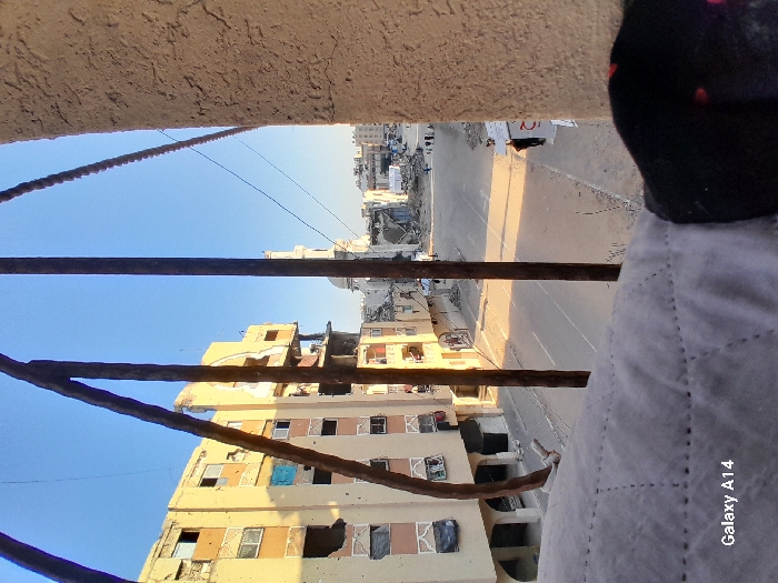 The image presents an elevated view of a street in Khan Yunis, Palestinian Territory, displaying significant urban destruction under a clear blue sky.
In the immediate foreground, three heavily corroded, dark metal bars run vertically, partially obscuring the view. To the right, a segment of a textured, light-colored stucco wall is visible. Below the bars, a section of light grey, quilted fabric occupies the lower left, with a small portion of a dark garment featuring red polka dots visible on the far right. A "Galaxy A14" watermark is present in the lower left corner.
The central focus is a two-lane paved road, appearing mostly empty but covered with debris, including rubble and dirt. Long shadows extend across the street from the left, indicative of either morning or late afternoon sunlight. On the left side of the street, a multi-story building with a light-colored facade shows substantial damage, including broken windows, missing sections, and some openings covered by dark material. Further down the street on the right, another multi-story structure also exhibits visible damage.
Several small human figures, approximately six, are discernible walking or standing in the distance on the right side of the street. Closer to the foreground on the right, a single individual in dark clothing is walking along the street's edge.
The background reveals extensive devastation across the urban landscape, with numerous buildings in varying states of collapse or severe structural damage. A distinctive white dome and a slender, white minaret, consistent with a mosque, rise above the damaged buildings in the mid-distance to the right of the frame, serving as a prominent landmark amidst the destruction. The overall environment suggests a scene of widespread conflict or disaster.