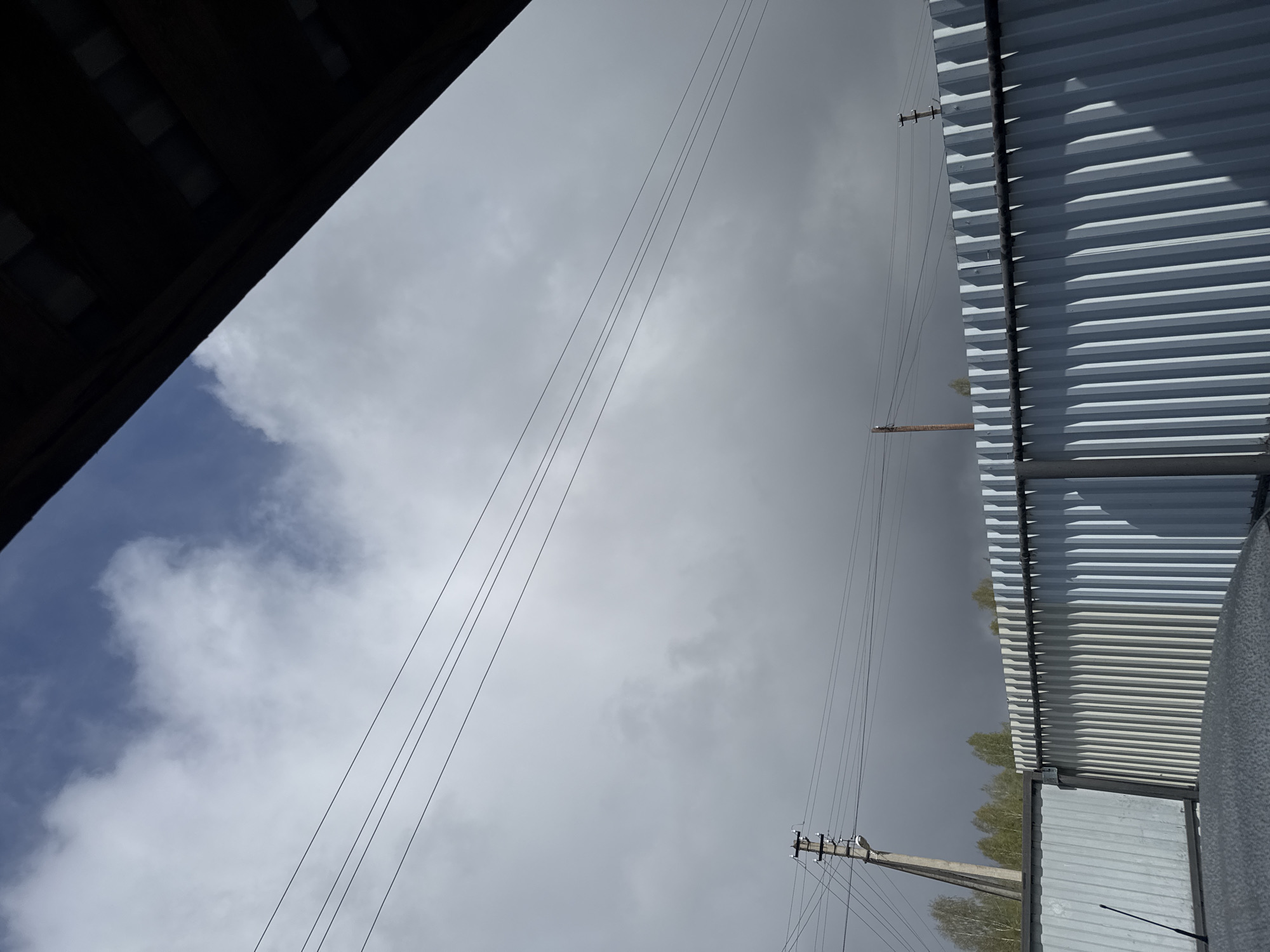 A low-angle shot captures a view toward the sky, with objects and structures in the foreground. Above is a sky filled with gray clouds, some areas show patches of blue sky. Power lines stretch across the image. The left portion of the image shows a dark building structure. The right part reveals metal corrugated structures, possibly rooftops or walls. Trees can be seen behind the metal sheet. The location is Borodulino, Russia, but further details are indiscernible from the given image.