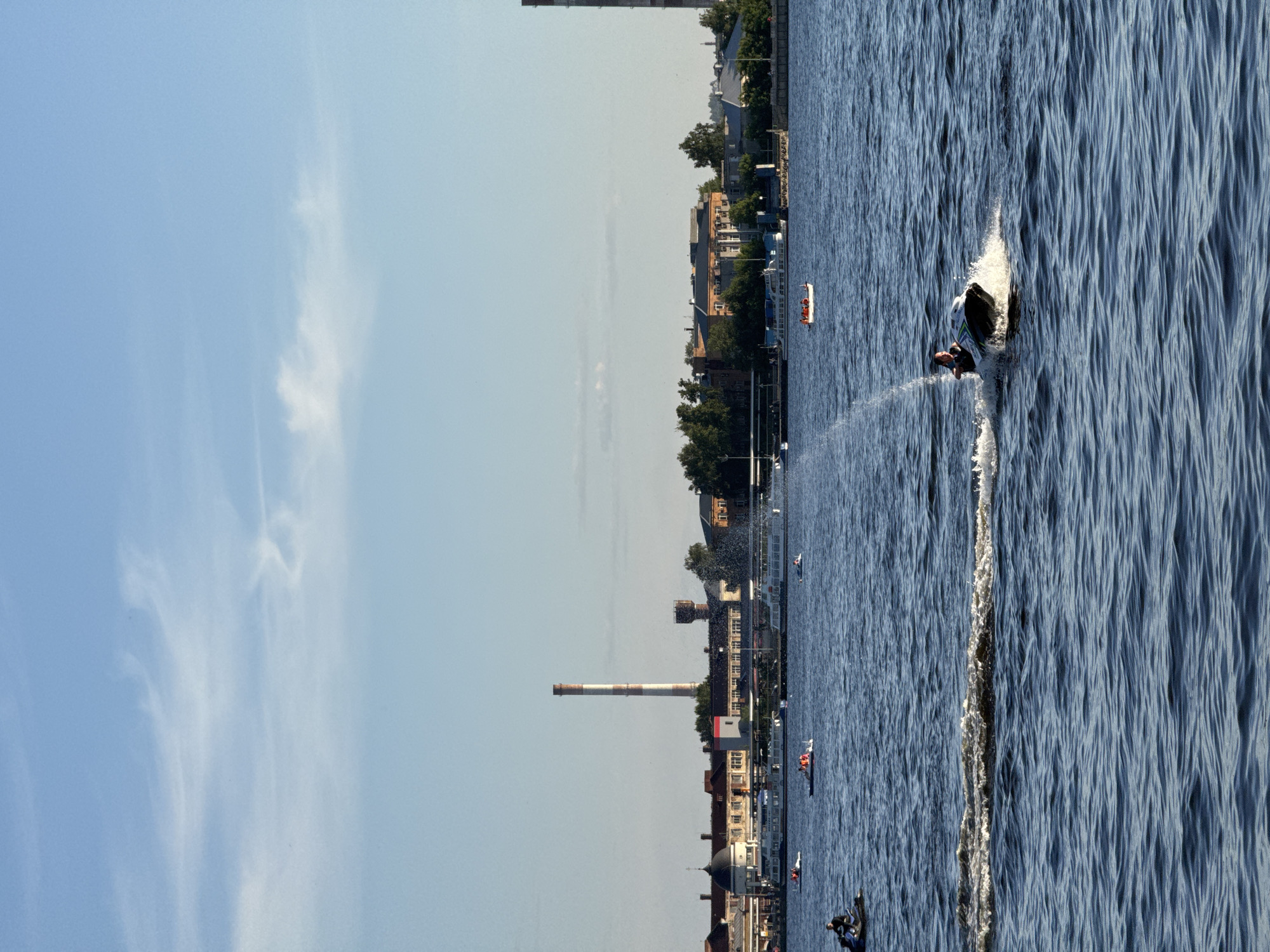 The image depicts a daytime scene over a large body of water in Izhevsk, Russia, likely the Izhevsk Pond or Reservoir, with a city skyline along the far bank. The sky, occupying the upper third of the frame, is clear light blue with sparse, wispy white clouds. The water, a dark blue with undulating ripples and small waves, dominates the middle and lower sections.
In the immediate foreground, to the lower right, an individual on a dark and white jet ski is navigating the water, generating a significant white spray from its rear and a distinct wake. Further back on the water, to the left, a person operates a smaller dark personal watercraft. Centrally, a small white boat carries multiple individuals, some visible wearing orange life vests. Another small white boat is positioned further right. Along the distant shoreline, several larger white passenger boats or ferries are docked.
The background cityscape features a prominent, tall industrial chimney with alternating red and white horizontal stripes, standing near the center-left. To the left of the chimney, various buildings with diverse architecture are visible, including older multi-story structures, one with a dark dome roof, and another featuring white pillars. To the right of the chimney, a dark water tower is discernible, along with additional buildings and green trees. A visible spray of water, resembling a fountain, rises from the water near the far bank on the right side. The scene is well-lit, indicating a sunny day.