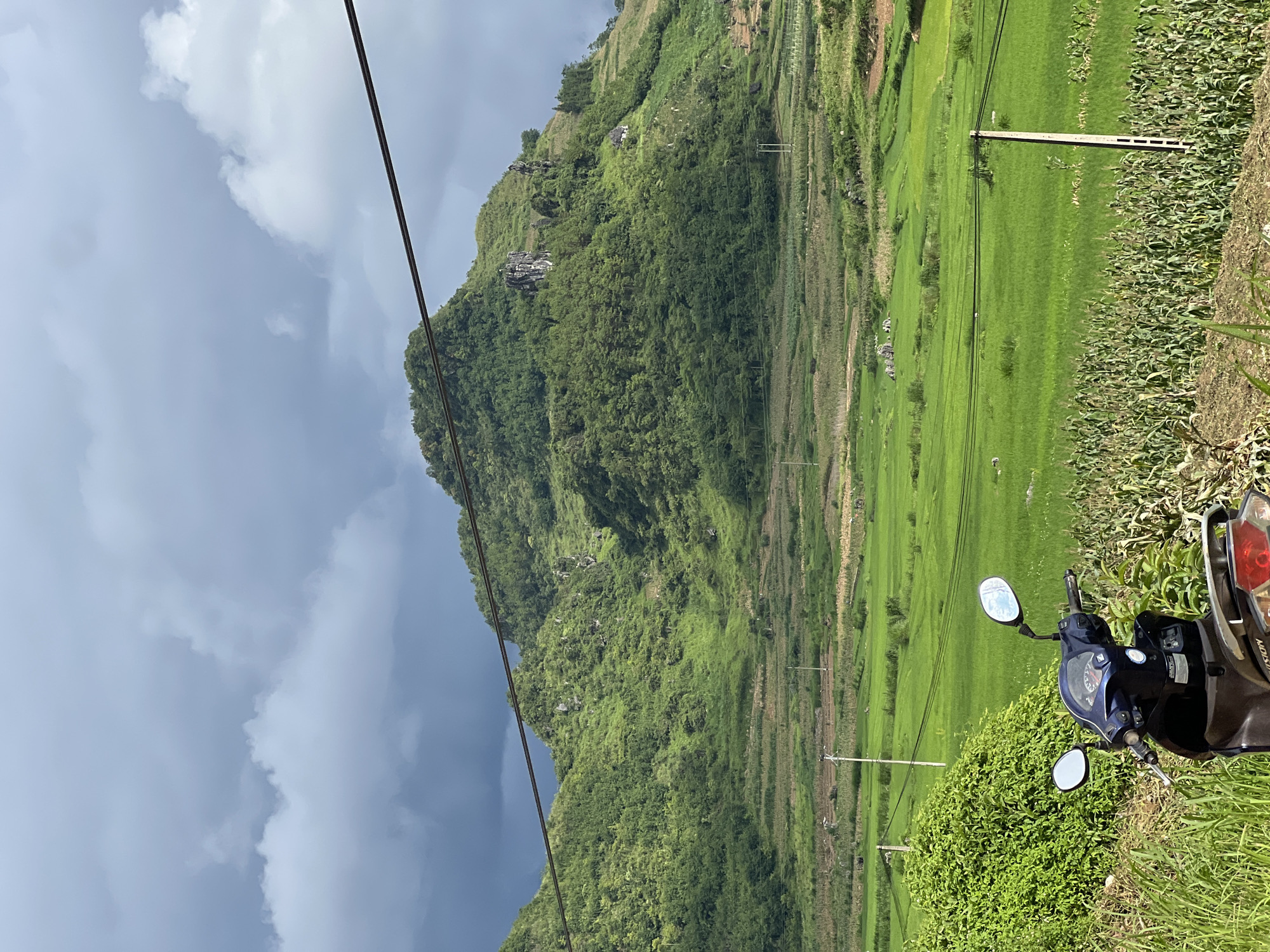 The image depicts a rural landscape in Quản Bạ Commune, Vietnam, under a dynamic sky. In the foreground, a dark blue scooter is parked amidst lush green vegetation and a field of corn. Behind the scooter, expansive, vibrant green rice paddies stretch across the middle ground, interspersed with terraced fields. A single utility pole stands to the right of the rice paddies. Dominating the background is a large, verdant mountain covered in dense trees, with some rocky outcrops visible on its slopes. The sky above is a mix of dark, moody clouds and brighter, diffused light. The overall scene suggests a natural and agricultural setting.