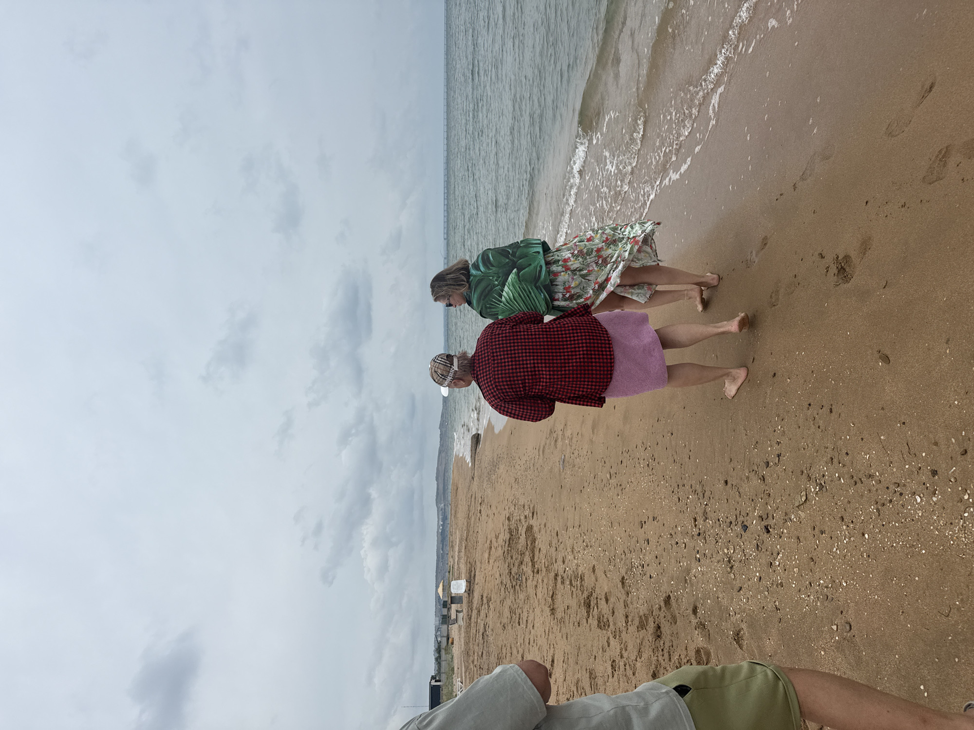 Three individuals are walking along a sandy beach. The person on the left, partially visible, is wearing a light green t-shirt and khaki shorts. The two individuals in the middle and to the right are walking away from the camera. The middle individual is wearing a red and black plaid shirt and a pink skirt. The individual to the right is wearing a green tropical-print garment and a floral skirt. The beach is composed of tan sand with scattered shells and pebbles. Gentle waves are lapping at the shoreline. In the distance, a line of buildings and a bridge are visible along the coast. The sky is overcast with gray clouds. The scene is consistent with a location on the Black Sea coast in Kerch, Ukraine.