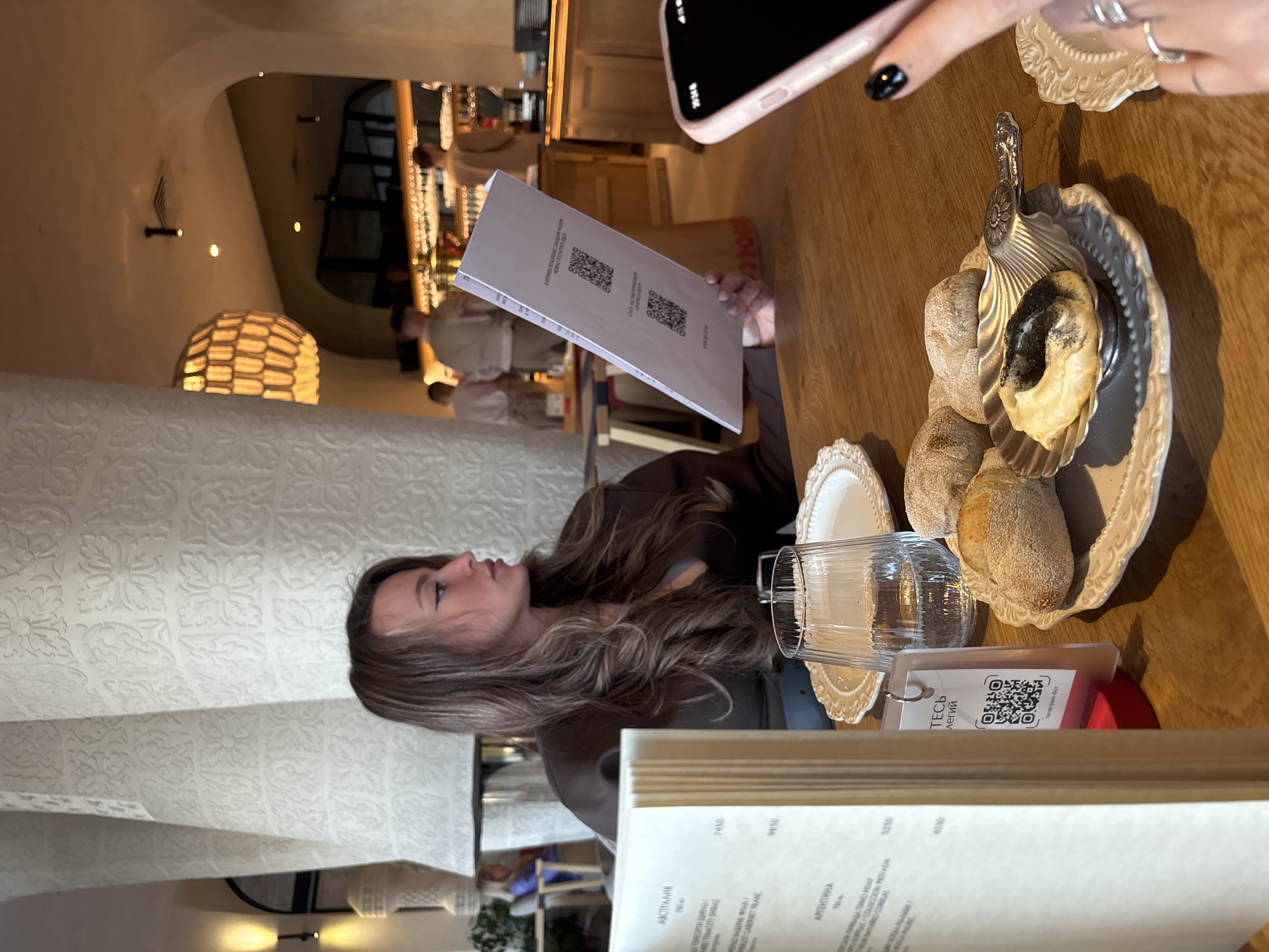 The image, taken inside a restaurant in Krasnoyarsk, Russia, shows a young woman with long brown hair looking at a menu. A partly visible cell phone is in the upper right corner. On the table are bread rolls on a decorative plate, a glass of water, and another menu with QR codes. The restaurant appears to be well-lit with textured walls and a view into a bar area. Other patrons are visible in the background.