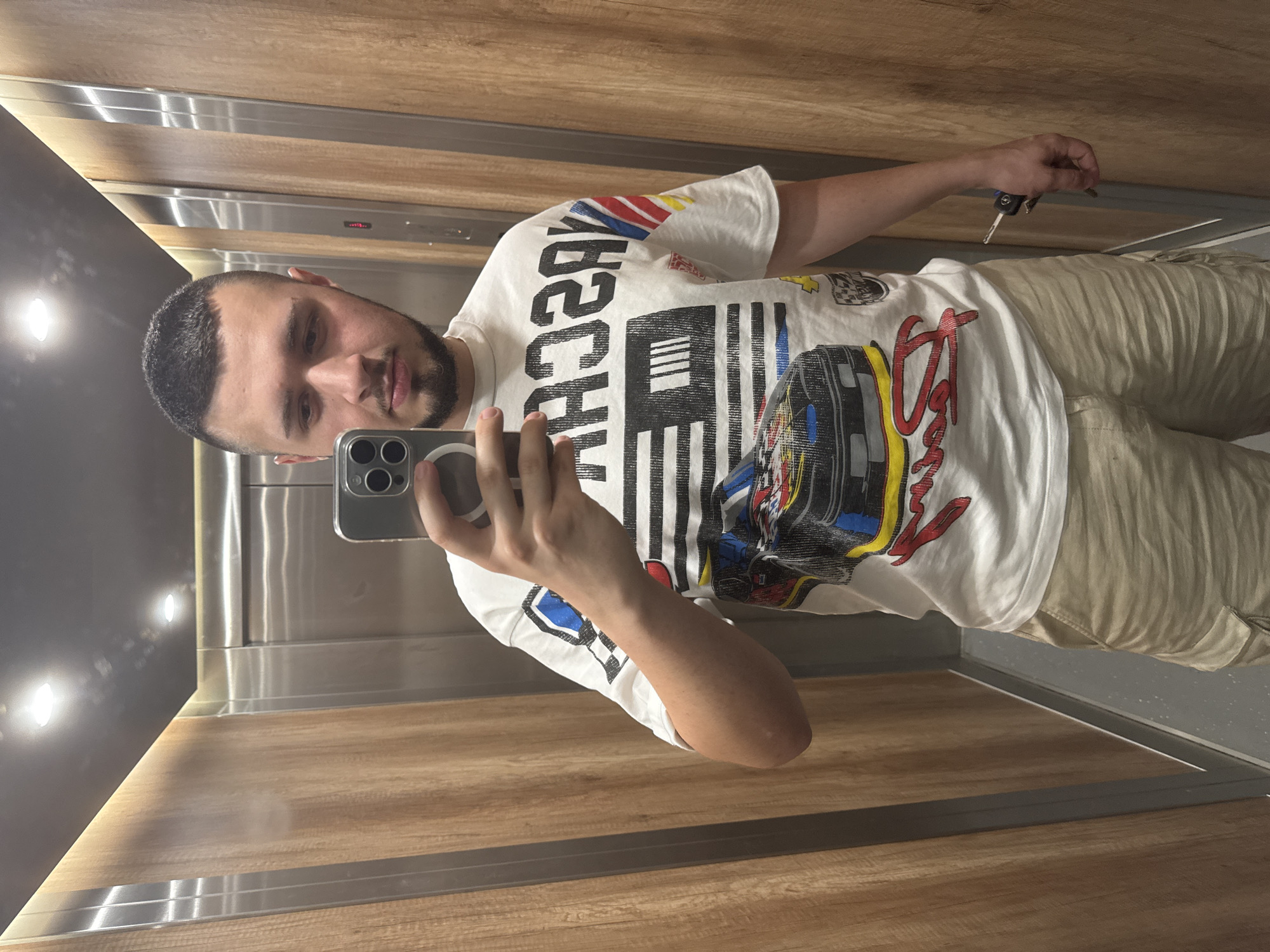 The image shows a young man with short hair and a beard taking a selfie in an elevator. He is wearing a white t-shirt with a graphic print of a race car and the word "NASCAR" on it, and khaki cargo shorts. He is holding his phone in his right hand, and a set of keys in his left hand. The elevator has wooden-paneled walls and stainless steel accents. The location is an elevator in Sofia, Bulgaria.