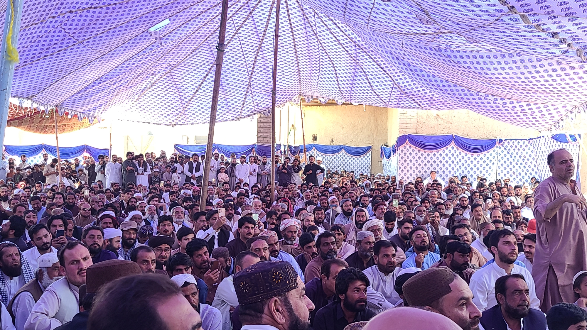 A large crowd of men is gathered outdoors under a large, patterned tent in what appears to be Panjpai, Pakistan. The crowd is densely packed, primarily composed of adult males with various traditional and Western attire. The environment suggests a formal or public gathering, possibly a religious or community event, based on the size and composition of the audience. The tent's draping and surrounding architectural features indicate a specific location within the city.