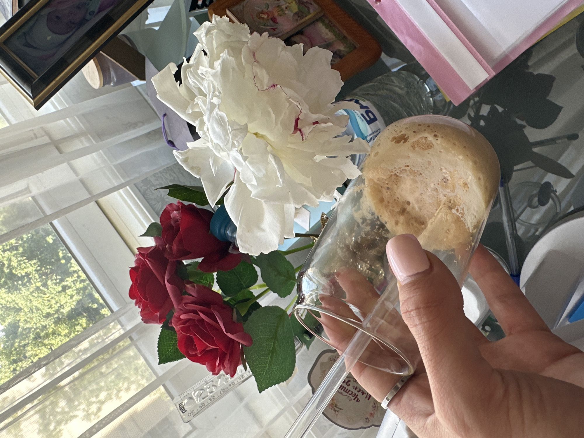 A close-up shot captures a person's hand, adorned with a silver ring and a light pink nail, holding a clear glass containing a frothy, light brown beverage. The glass is tilted, revealing the texture of the drink. In the background, a bouquet of red roses and a large white peony are arranged. A window with sheer white curtains allows natural light to illuminate the scene, with greenery visible outside. A framed picture and a water bottle are also present on a glass table. The setting appears to be an indoor domestic environment, likely a home or a cafe, with no specific identifiable landmarks indicating the city of Sofia, Bulgaria.